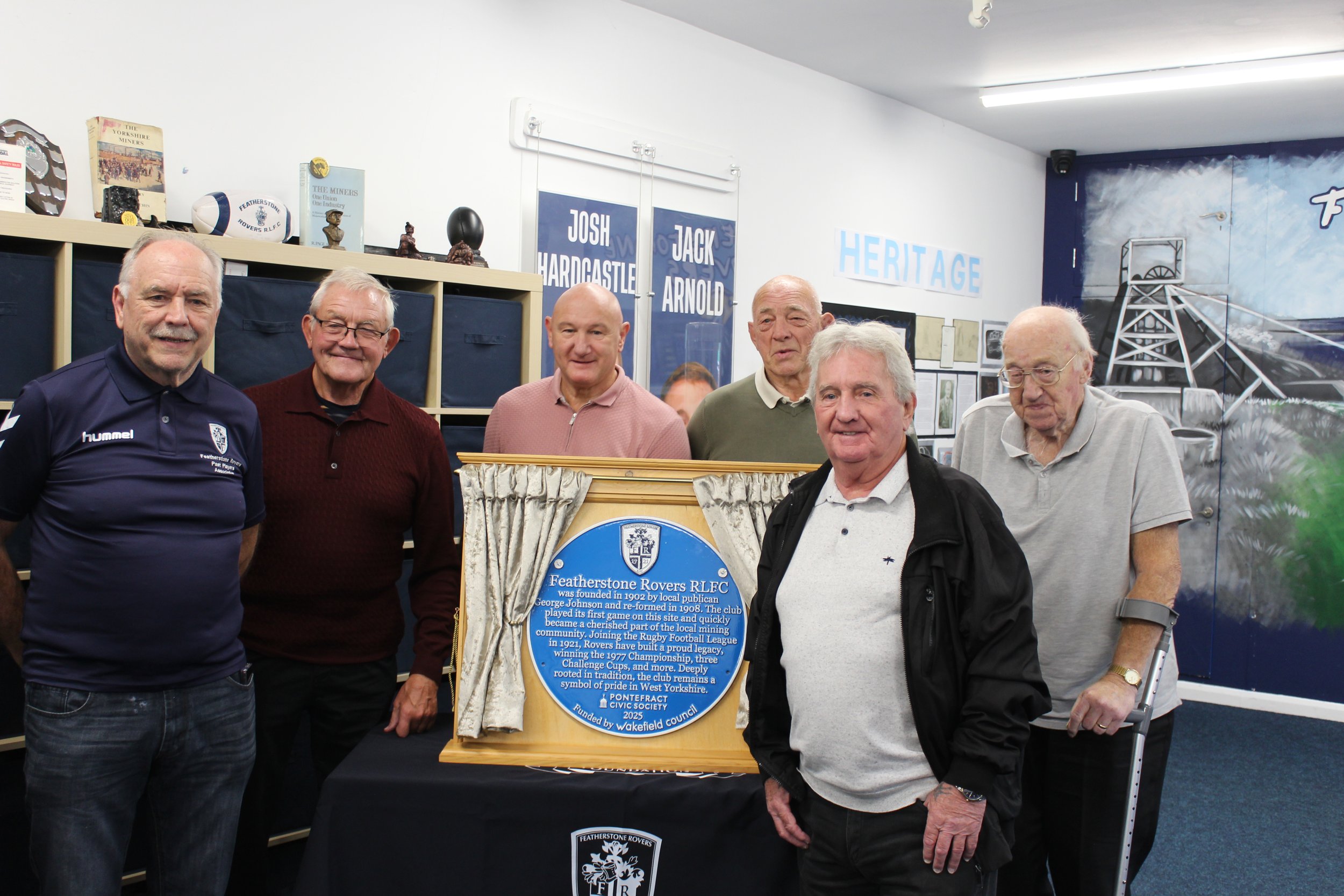 Six men standing behind a blue plaque about Featherstone Rovers RLFC, with a backdrop of sports memorabilia and a wall with blue and white decorations.