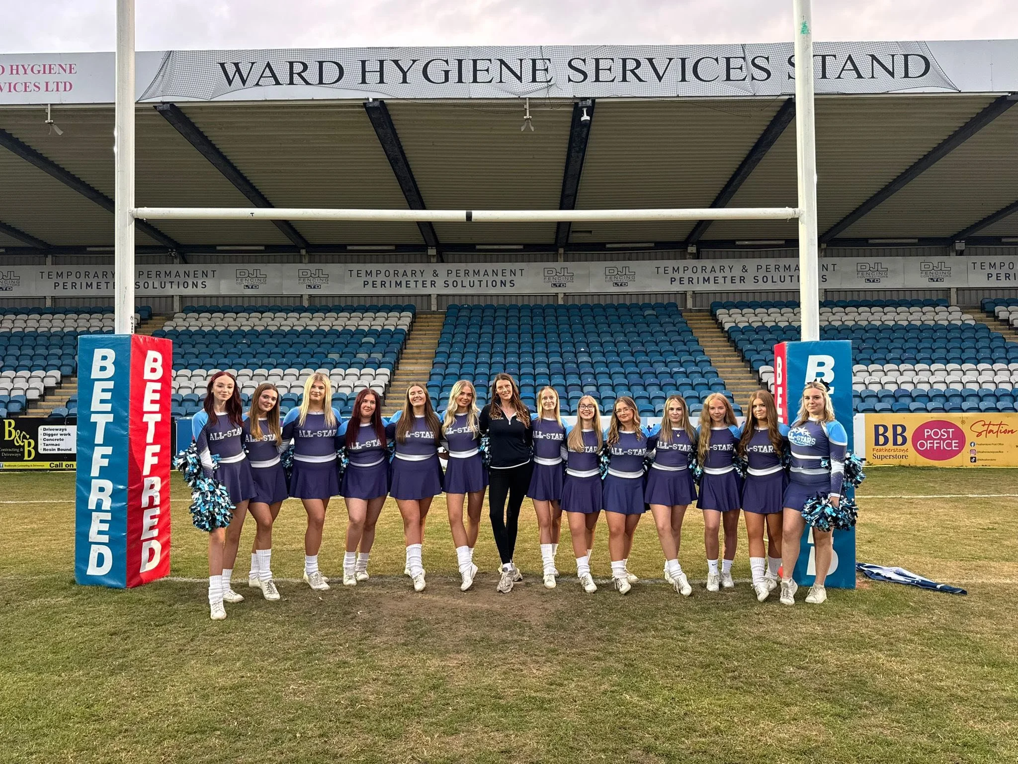 A group of cheerleaders in purple uniforms standing in front of a stadium, with a large banner that says 'Ward Hygiene Services Stand'.