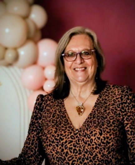 A smiling woman with shoulder-length hair wearing glasses, a leopard print top, and a heart-shaped pendant necklace, standing in front of pink and beige balloons on a pink background.