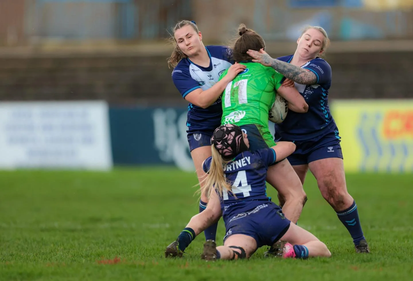 Three women, two in blue rugby uniforms and one in green, are tackling a woman in a green rugby uniform holding a ball during a rugby match on a grass field.