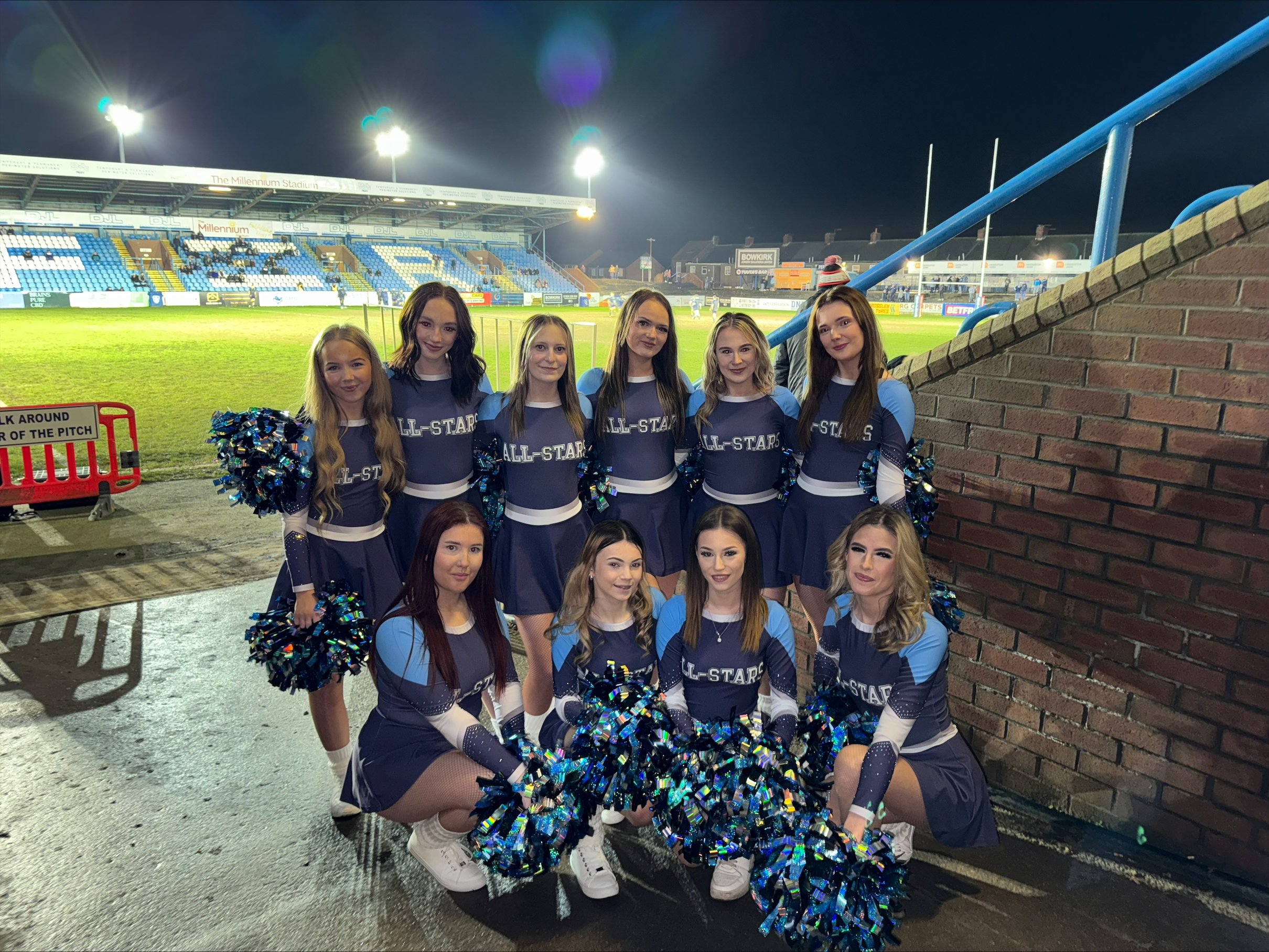 Group of cheerleaders in blue uniforms posing on a football field at night, with stadium lights in the background.
