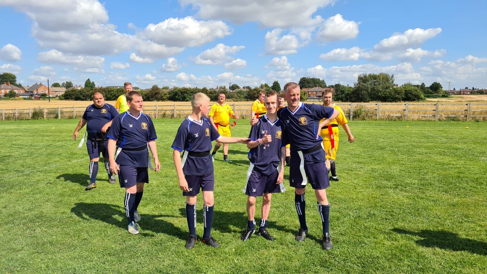 Group of people in sports uniforms walking on grass field on a sunny day with houses and trees in the background.