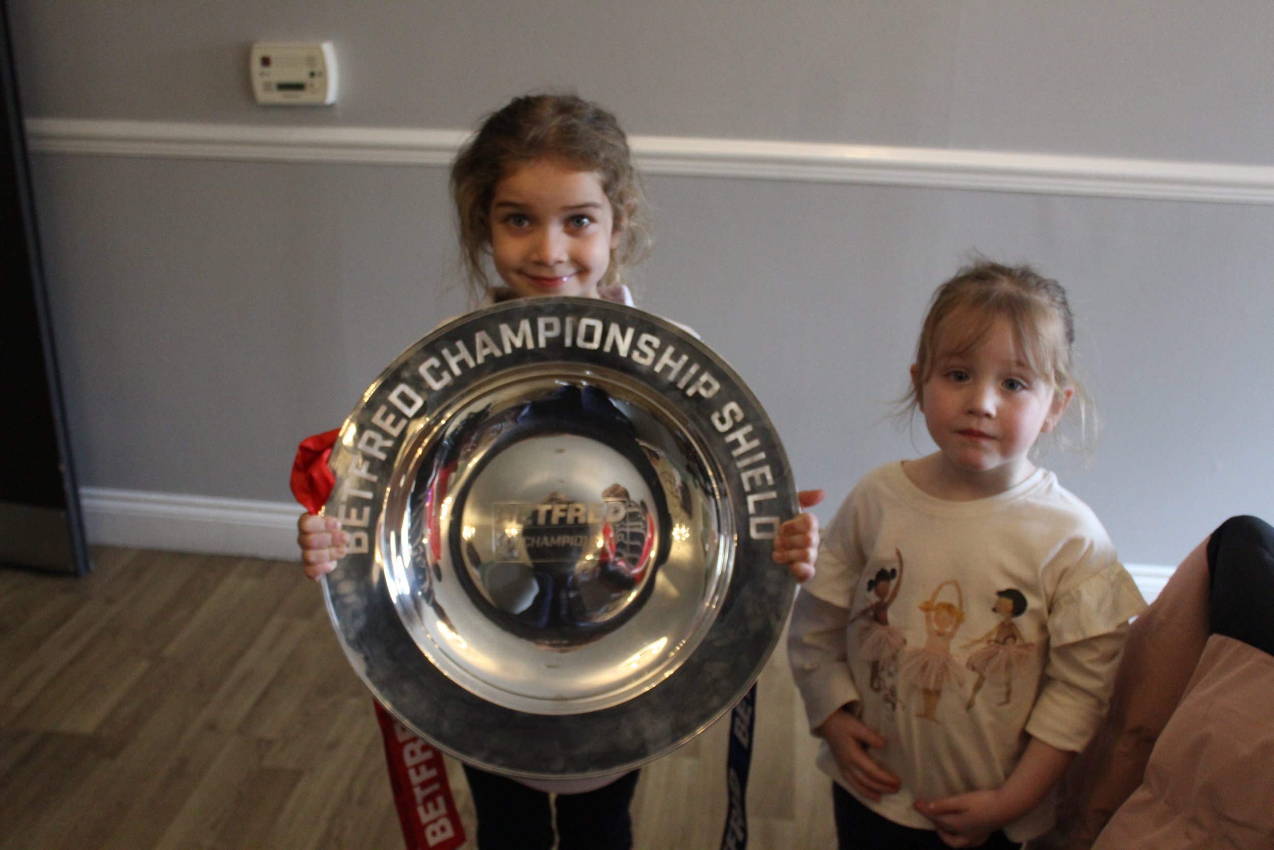 A girl holding a large hockey trophy with a younger girl standing beside her, both indoors in front of a gray wall.