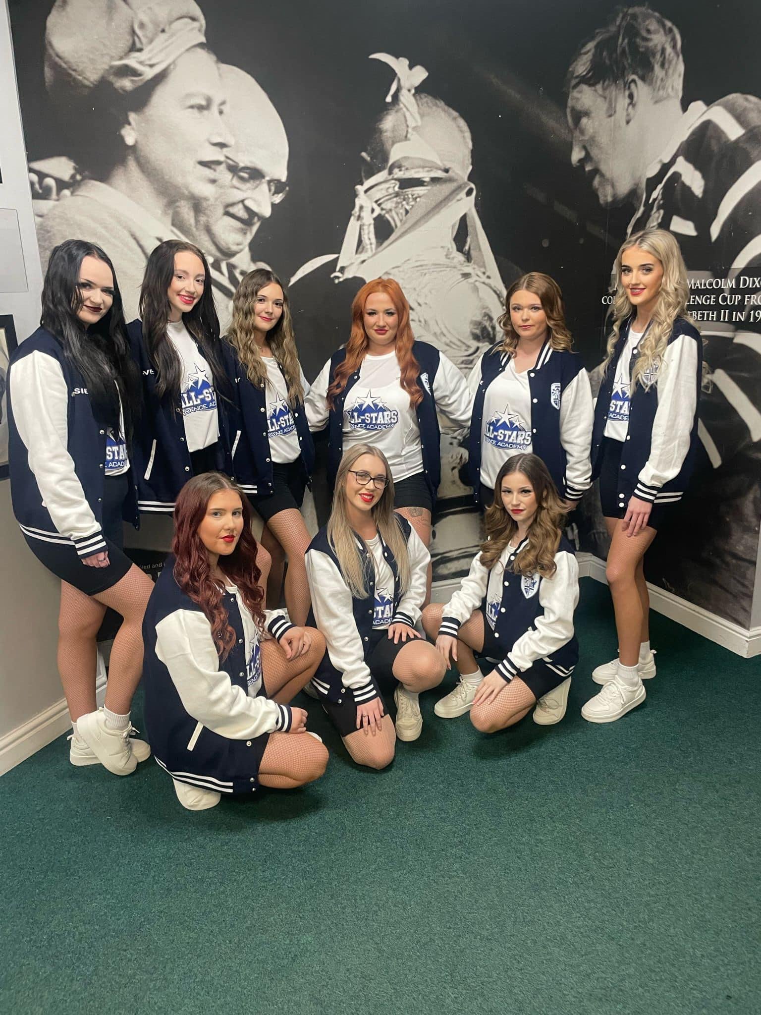 Group of young women dressed in cheerleading uniforms with 'All-Stars Dance Academy' logos, posing indoors with a large black-and-white photo of historical figures in the background.