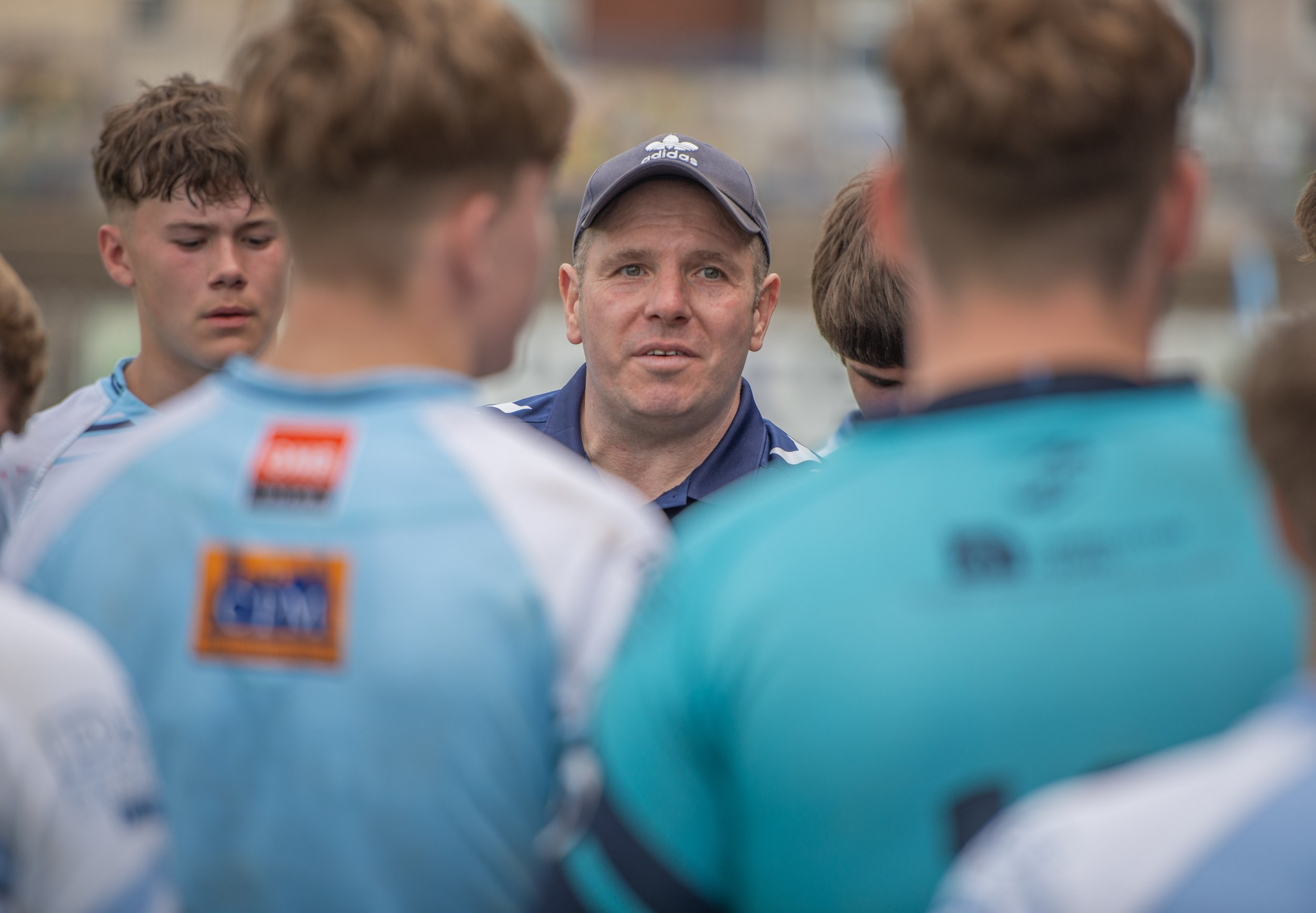 A man, likely a coach, speaking to a group of young male athletes in a sports setting. The coach is wearing a dark cap and shirt, and the athletes are dressed in sports jerseys, with some displaying patches and logos. The focus is on the coach's face, with the athletes' backs and faces blurred in the foreground.