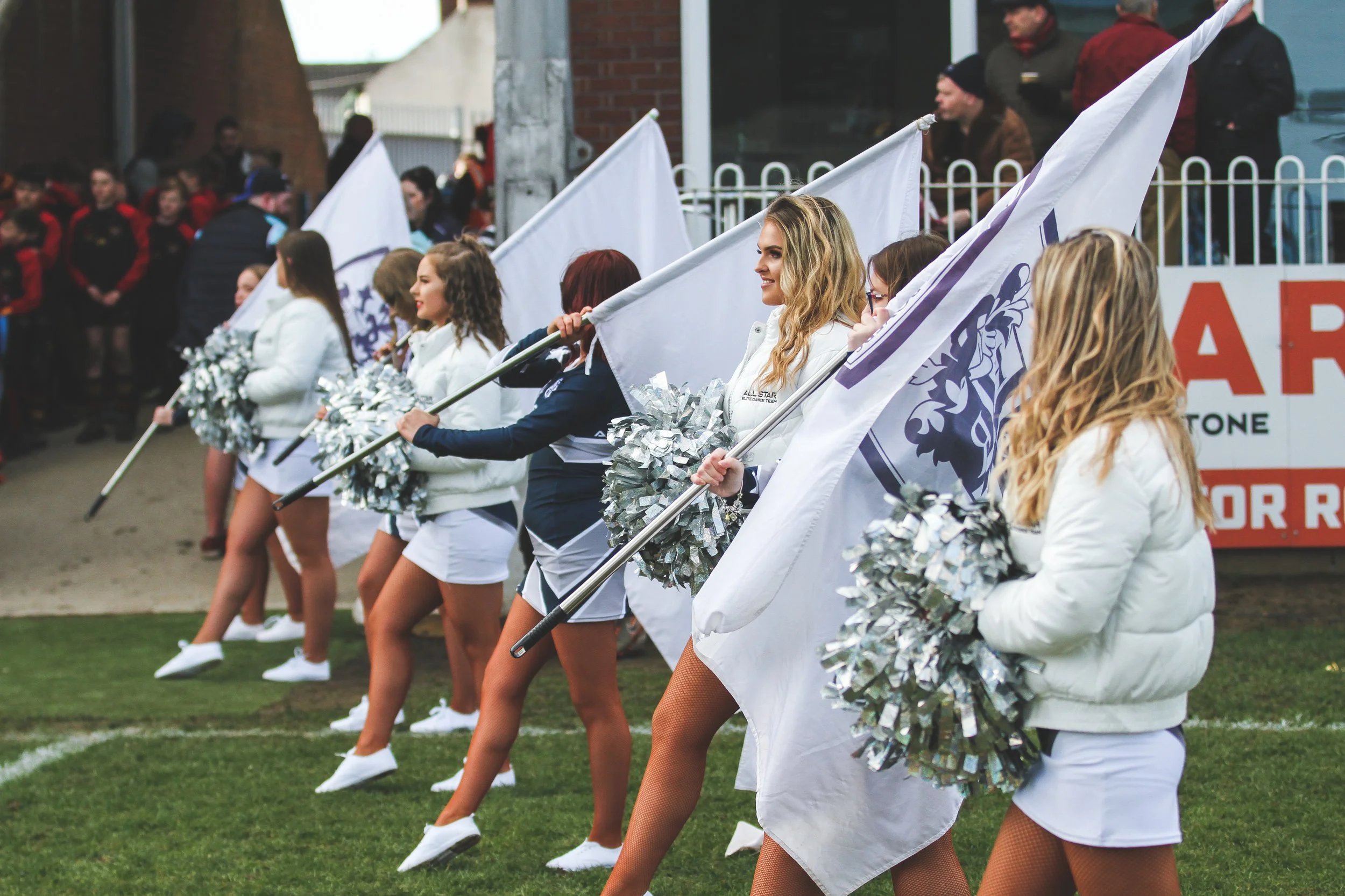 Women cheerleaders dressed in white jackets, holding silver pom-poms and flags, perform a dance routine on a field during an outdoor event, with a crowd in the background.