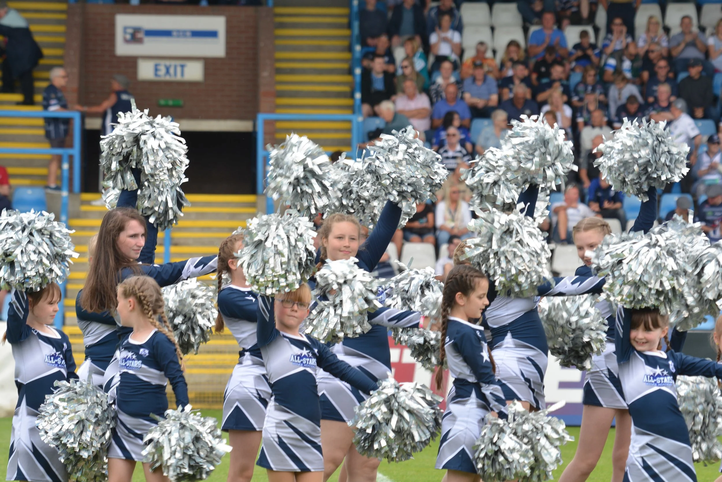 Group of young cheerleaders in navy and white uniforms performing with silver pom-poms on a football field with audience in the stands.