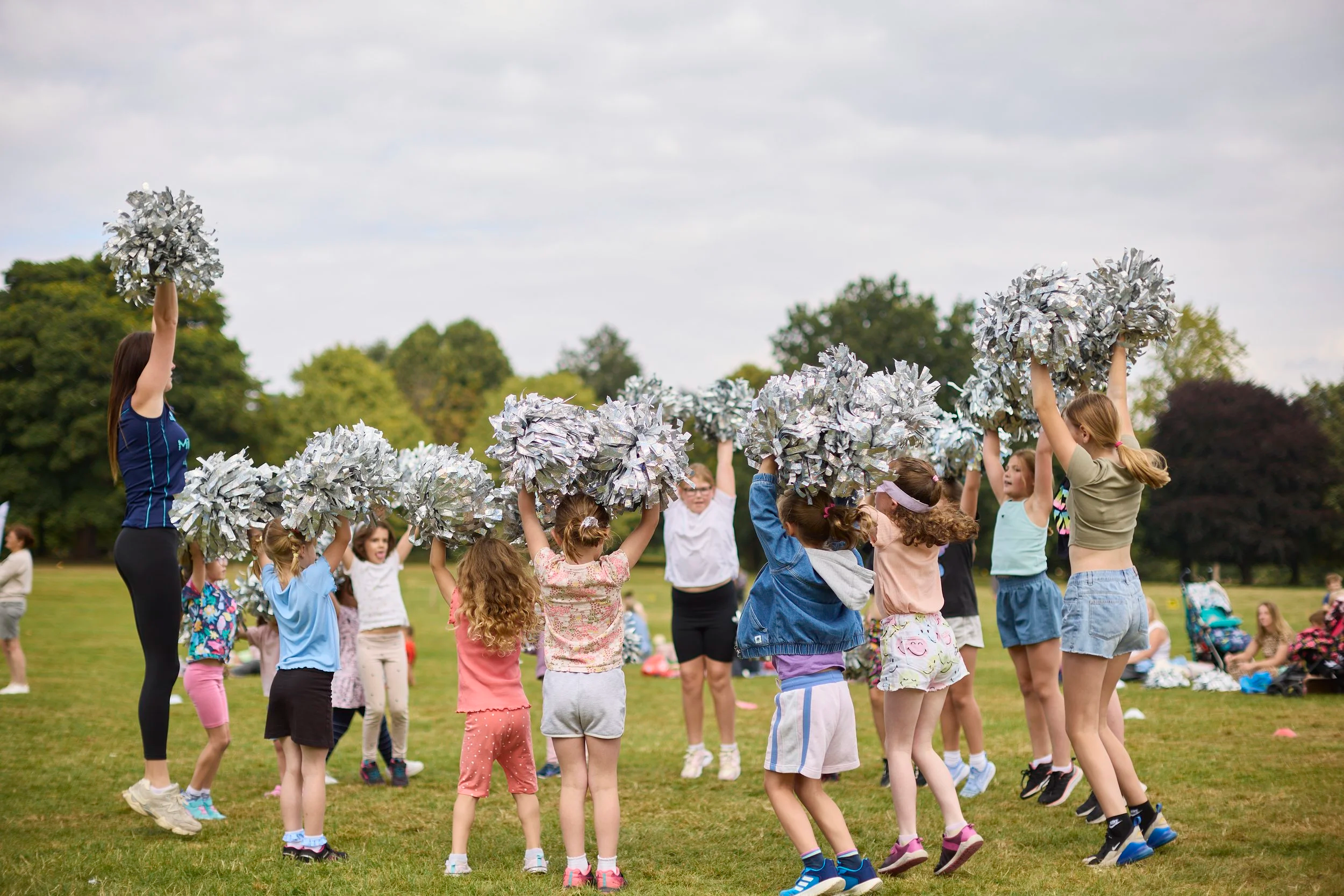 Children and cheerleaders in a park holding silver pom-poms during an outdoor cheerleading practice or event.