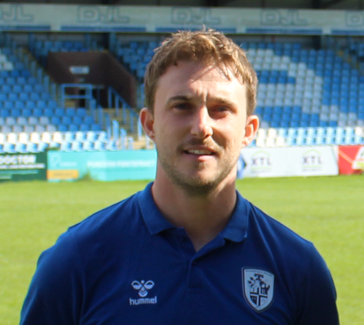A man with short fair hair wearing a blue sports polo with a blue and white design, standing against on a rugby pitch of grass.