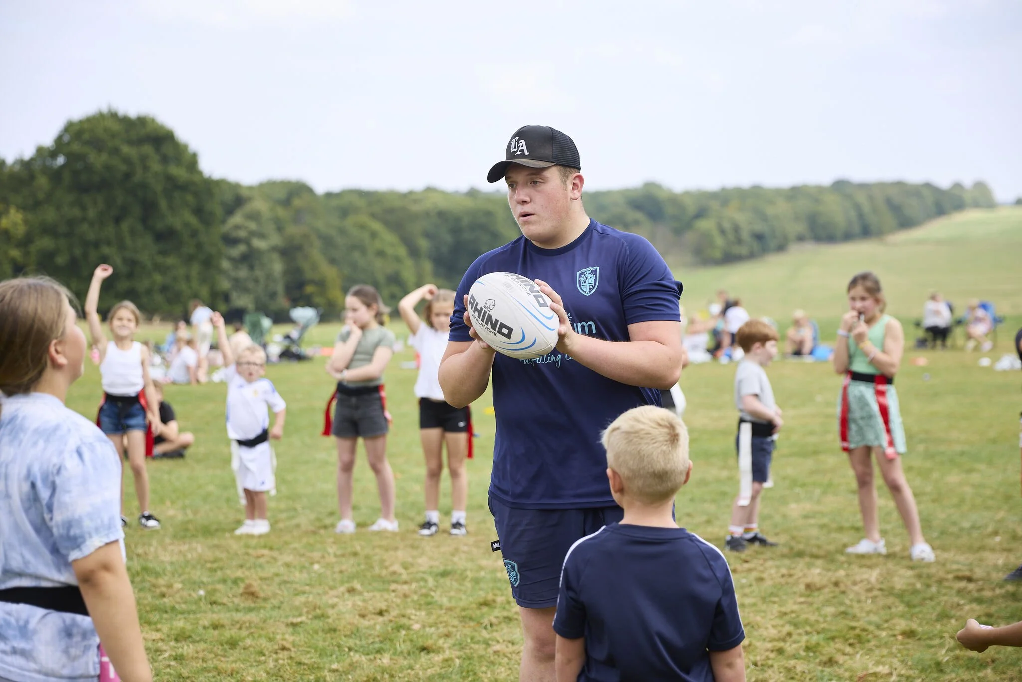 A young man with a black baseball cap and a navy blue sports jersey holding a rugby ball, surrounded by children outdoors on a grassy field with trees in the background.