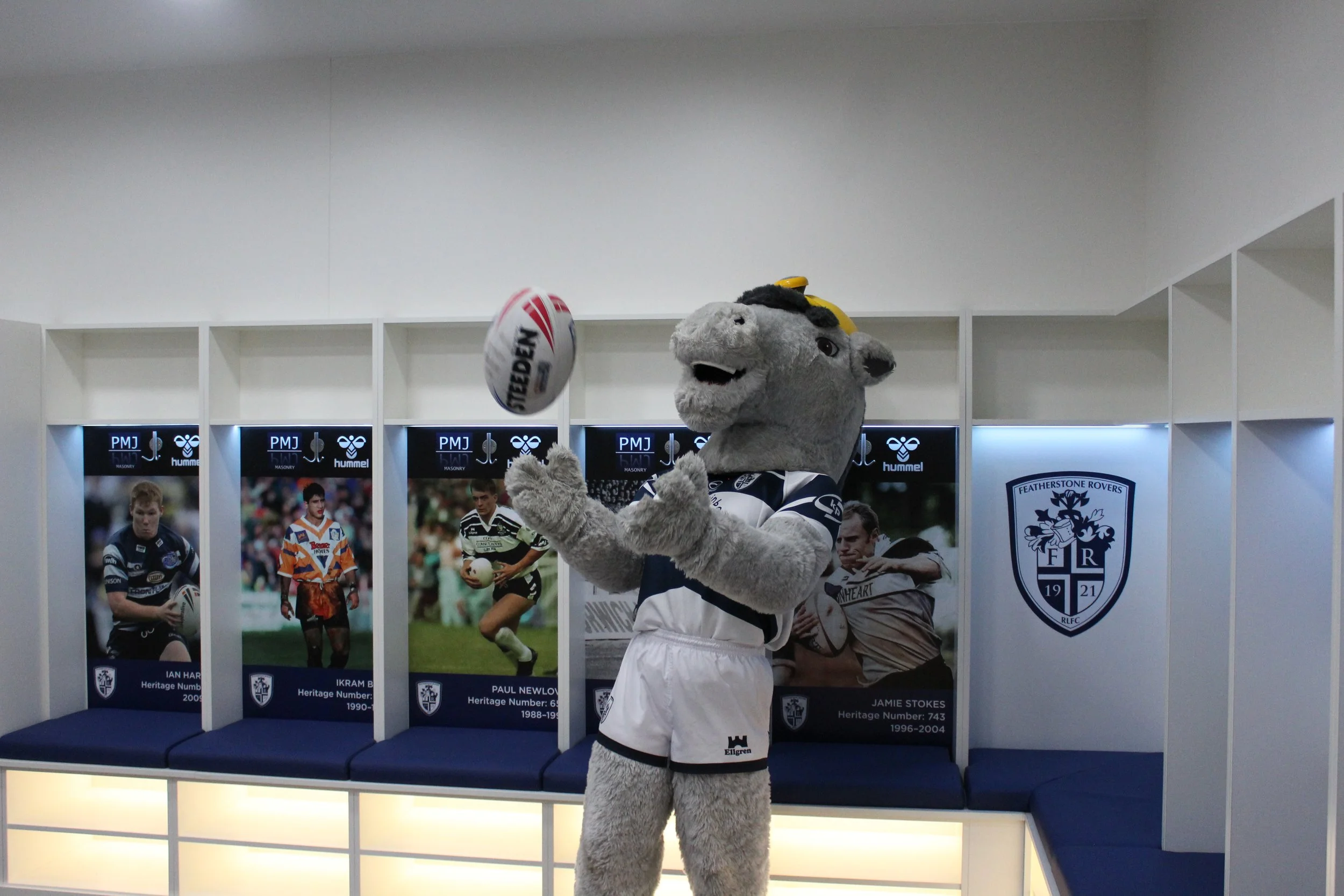 A mascot dressed as a wolf, wearing a football jersey, is throwing a rugby ball in front of a display featuring rugby player photographs and player information. The setting appears to be indoors, possibly at a rugby club or event.