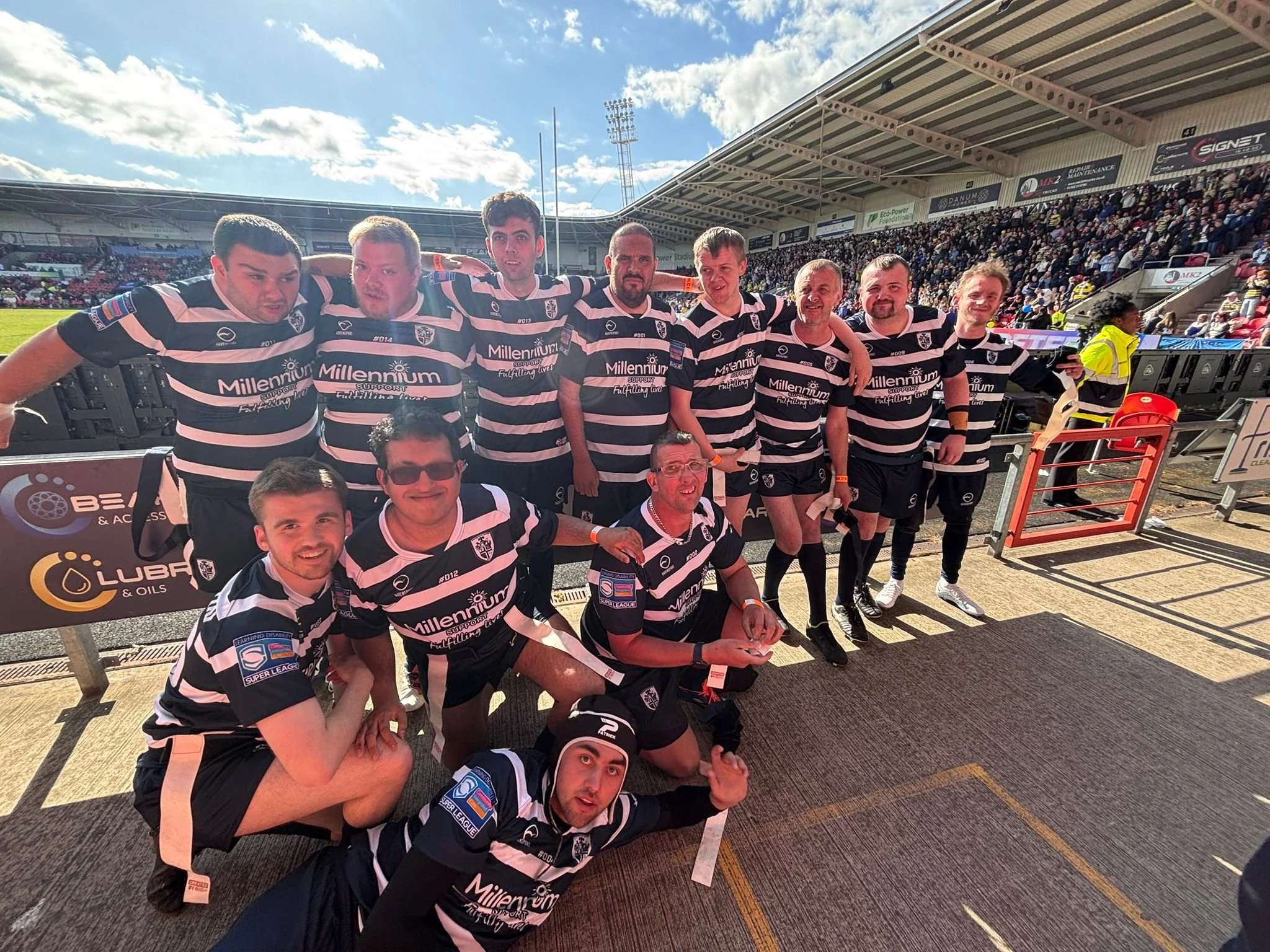 Group of rugby players in black and white striped jerseys posing together on a rugby field with a stadium full of spectators behind them.