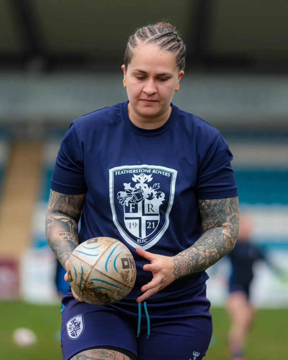Female athlete with braided hair and tattoos on arms, wearing blue sports uniform and holding a rugby ball during practice or game.