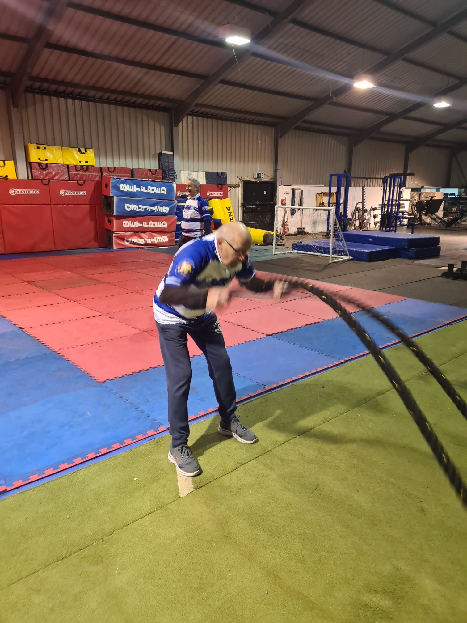 Indoor sports facility with padded flooring, two men are engaged in a fitness activity with battle ropes, one man is in the foreground and another is in the background near padded mats, gym equipment and mats are visible in the background.