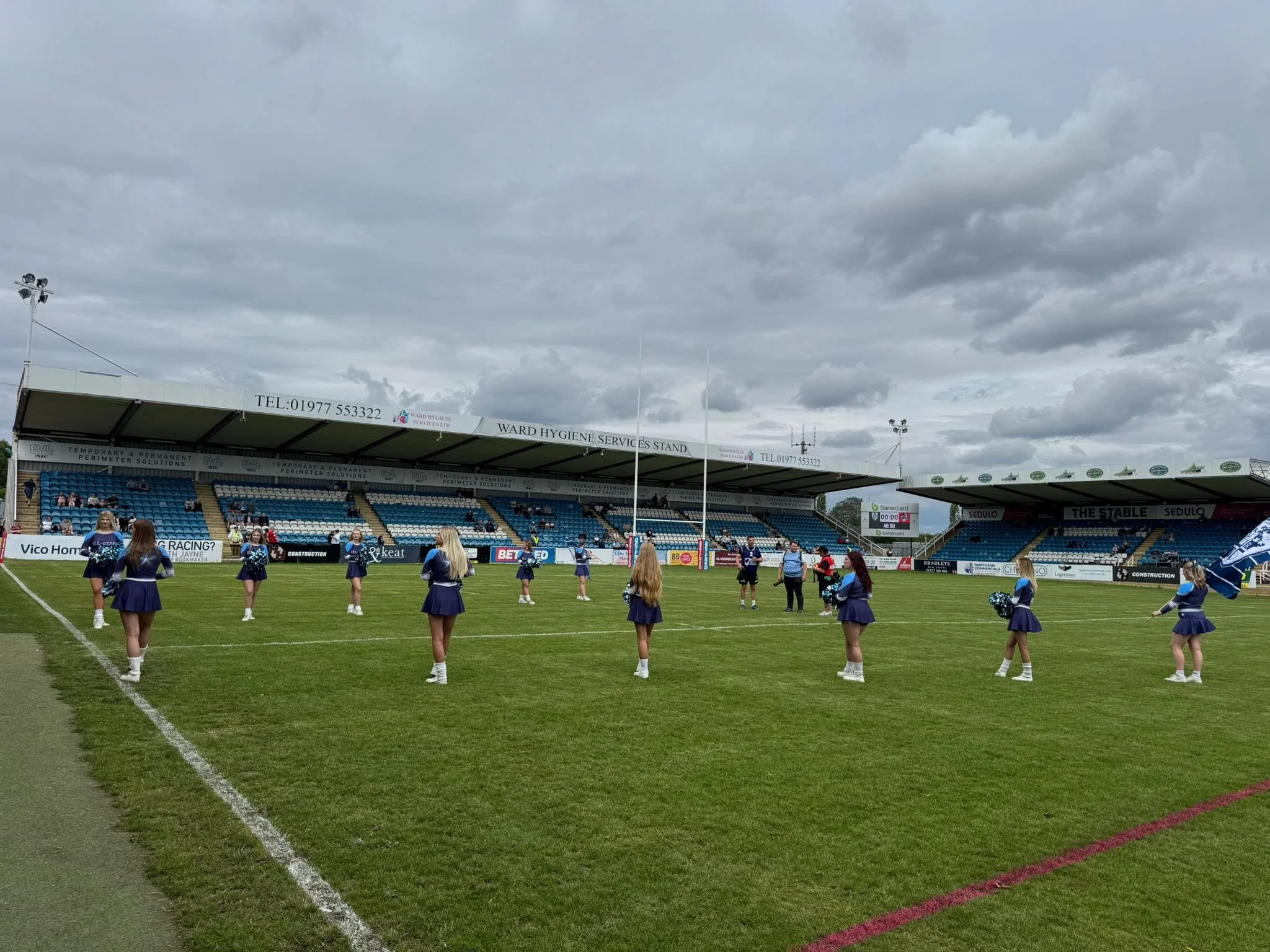 Cheerleaders practicing on a sports field before a game with stands and cloudy sky in the background.