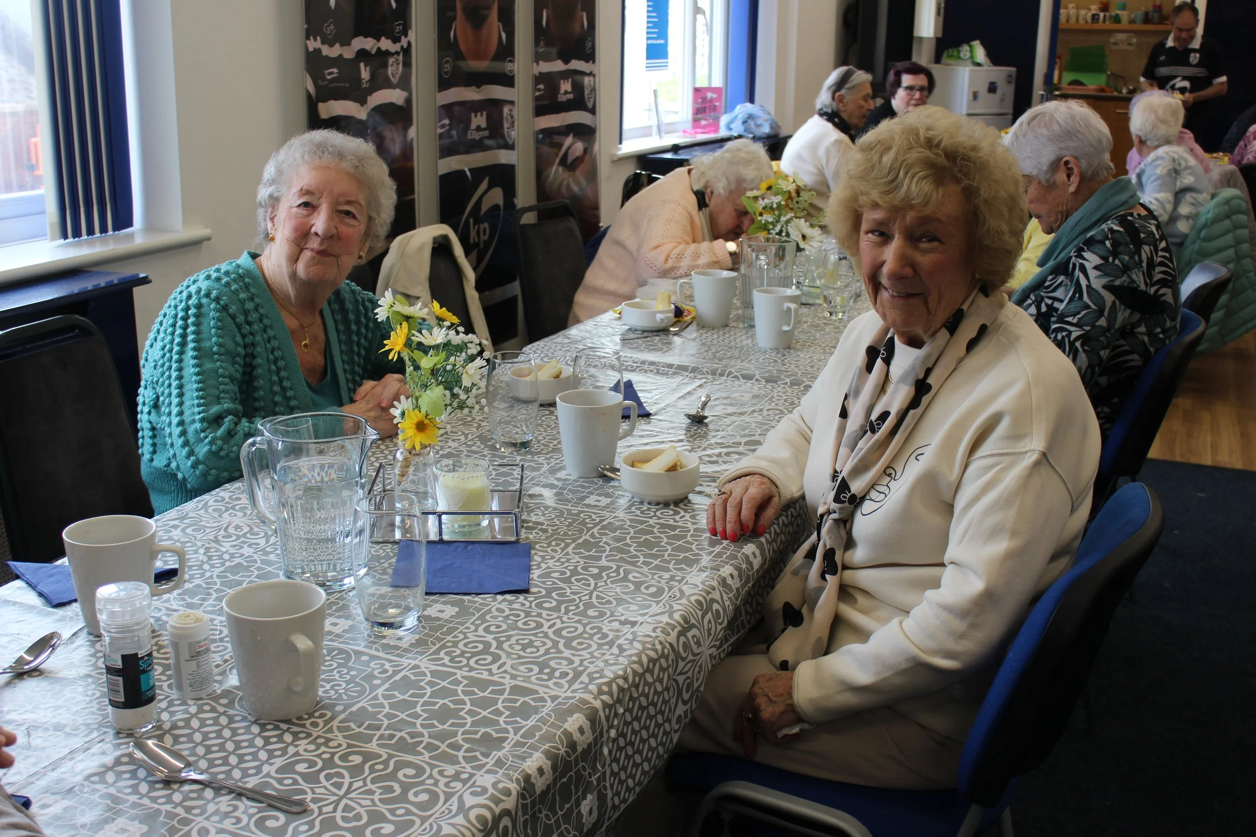 Group of elderly women sitting around a table with coffee cups and flowers during a social gathering in a well-lit room.