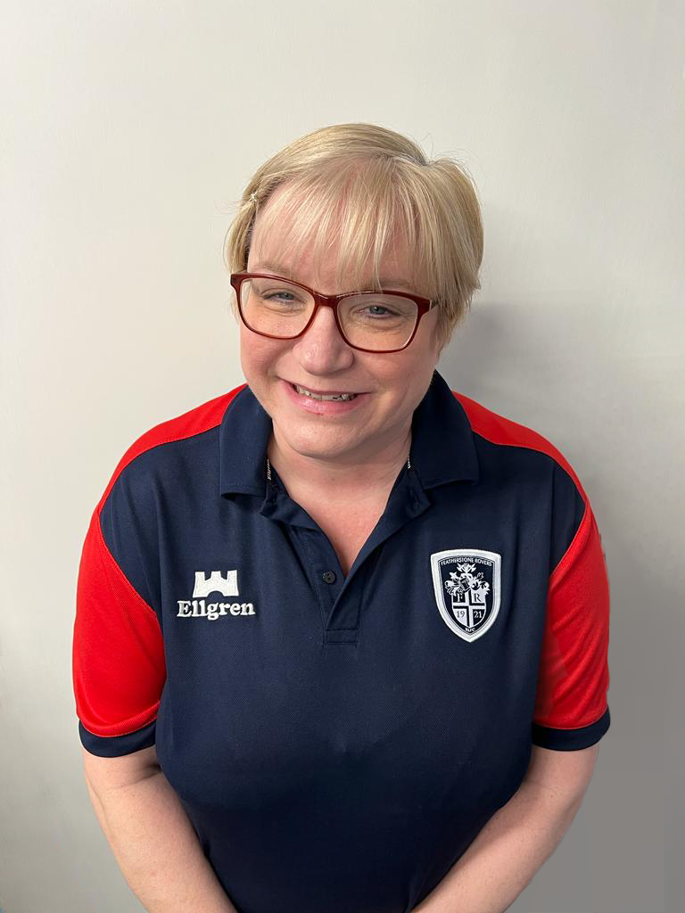 A woman with short blonde hair and glasses smiling, wearing a navy blue and red sports shirt with the logo 'Ellgren' and a crest patch.