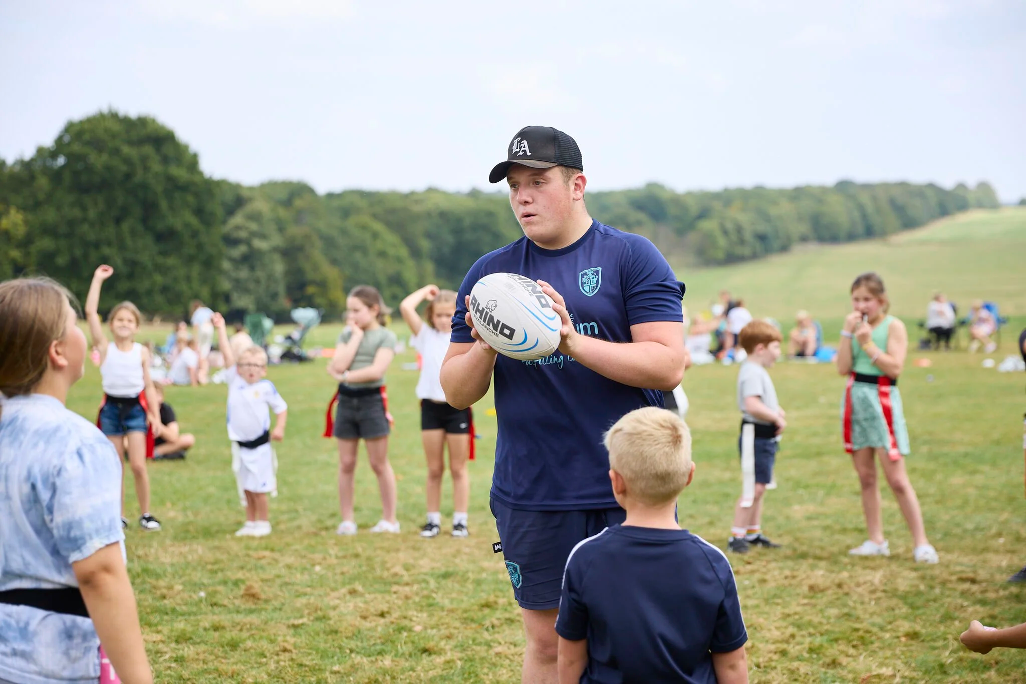 A young man in a navy blue shirt and black cap holding a rugby ball during a children's sports event on a grassy field with children and parents in the background.