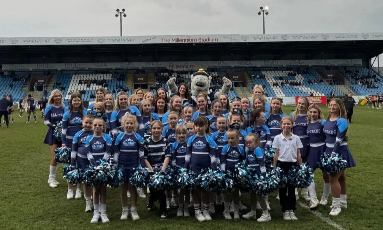 Group of cheerleaders and a mascot standing on a football field with stadium seats and a scoreboard in the background.