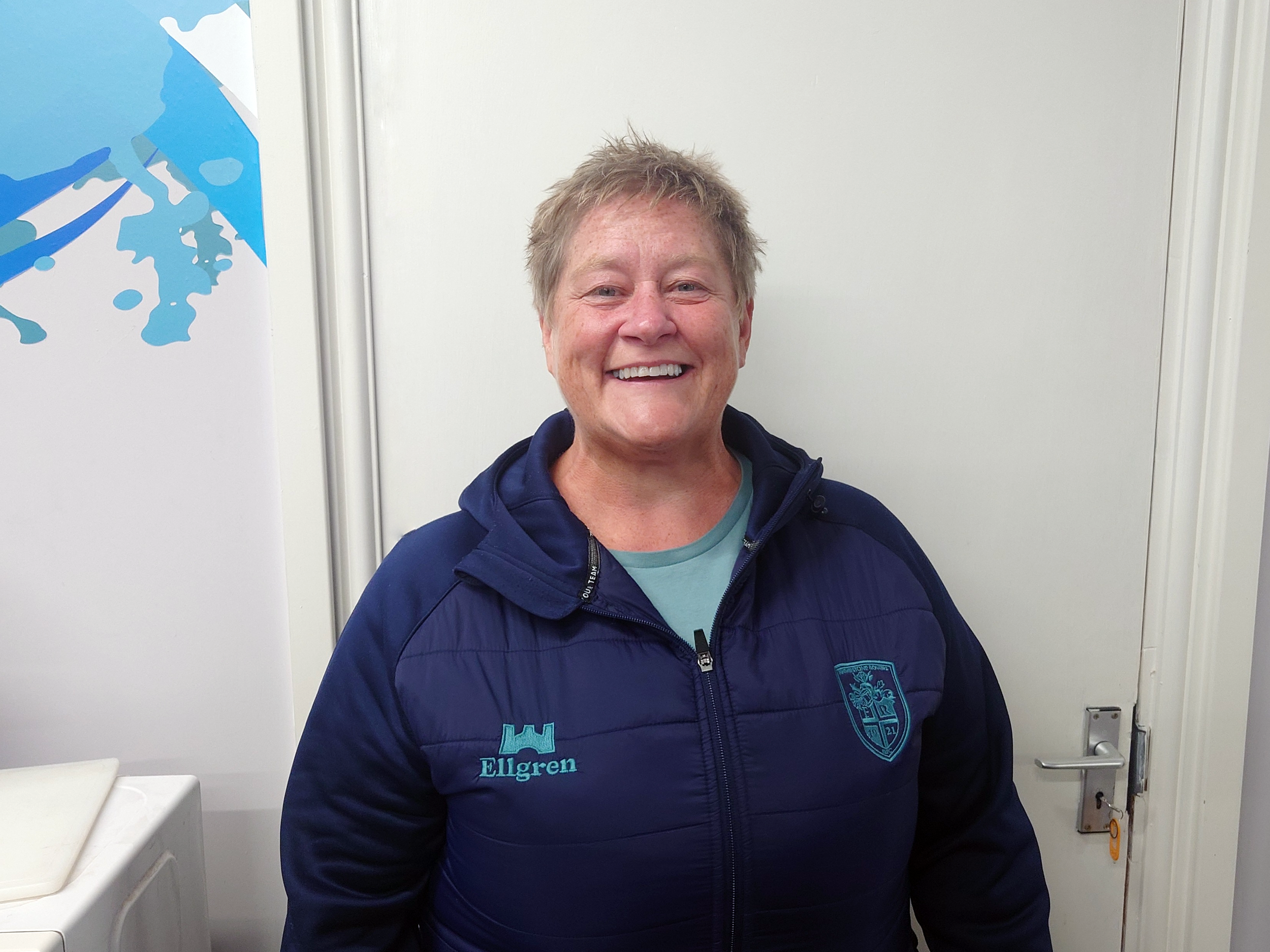 A smiling woman standing in a laundry room next to a washing machine and dryer. She is wearing a blue jacket with an embroidered emblem and the word 'Elgret' on it.