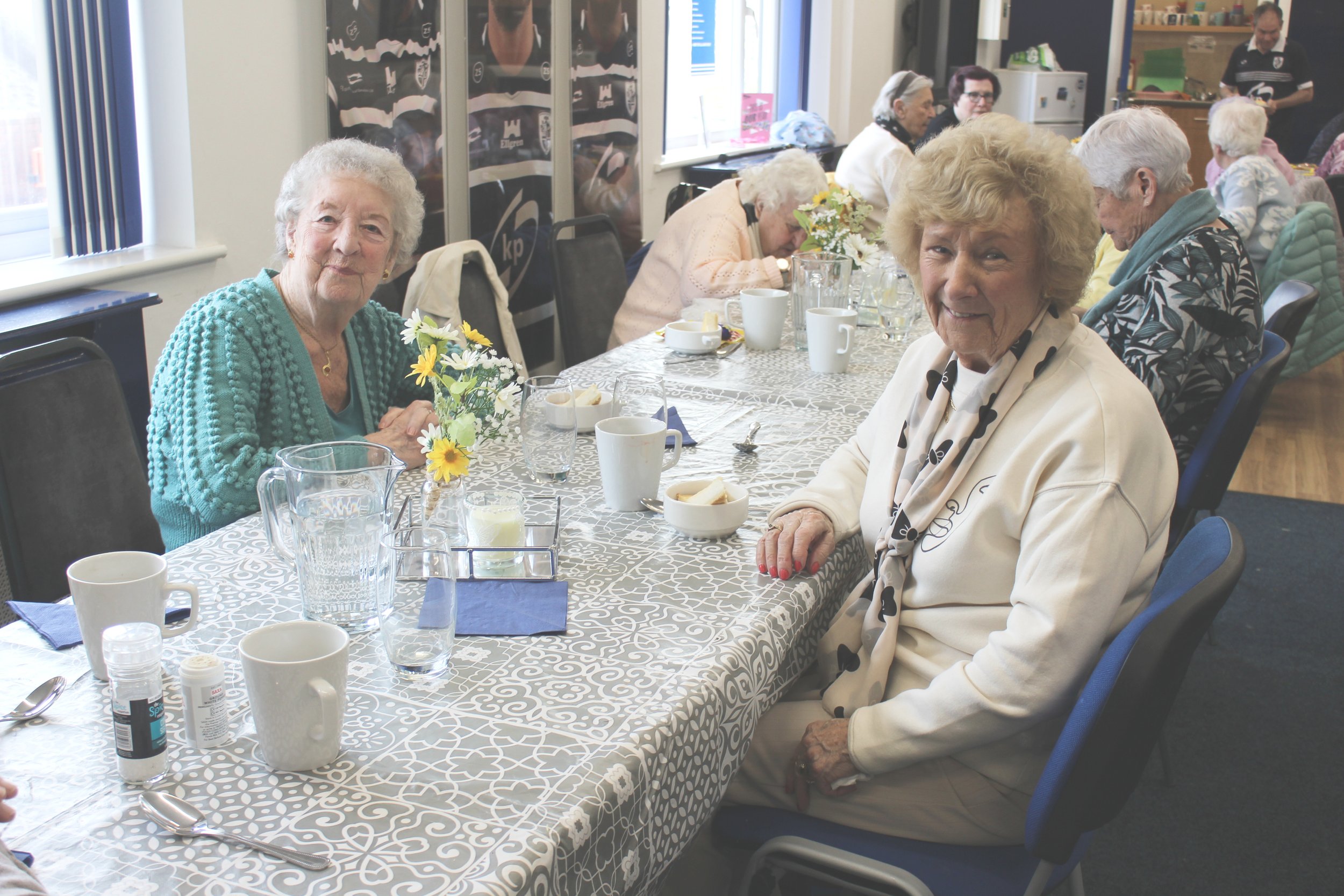 Older women sitting at a long table set for a meal, with cups, plates, and flowers, in a dining room with large windows and some people in the background.