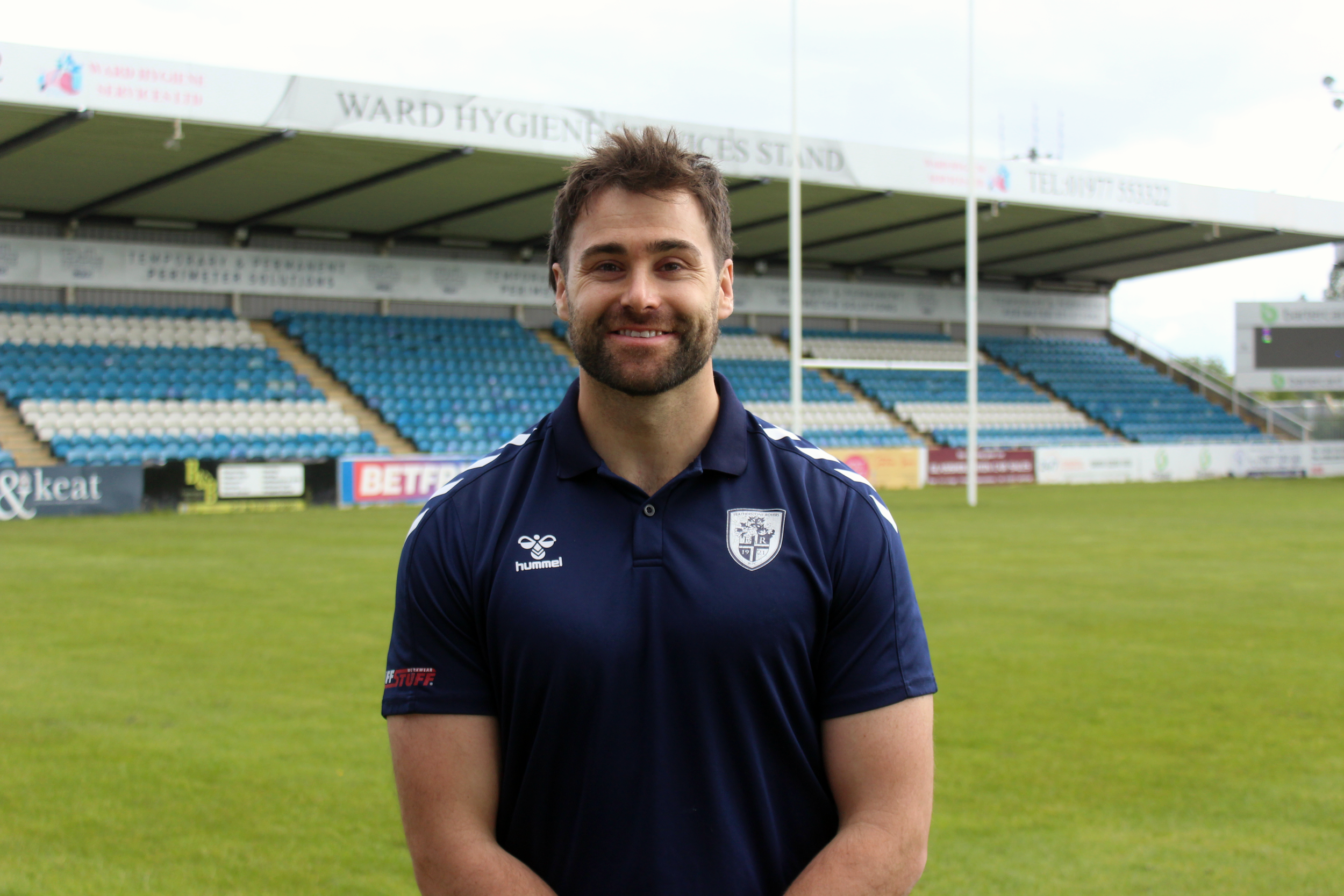 A man wearing a dark blue polo shirt and black pants standing next to a featherstone rovers foundation banner. The banner features the Featherstone Rovers mascot, a stuffed animal, and the foundation's logo.