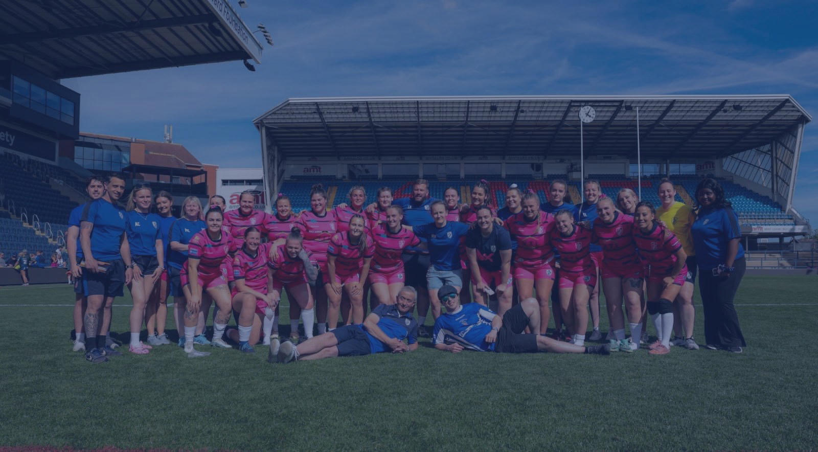 A group of women athletes in pink and blue sports uniforms with coaches, posing for a team photo on a sports field at a stadium.