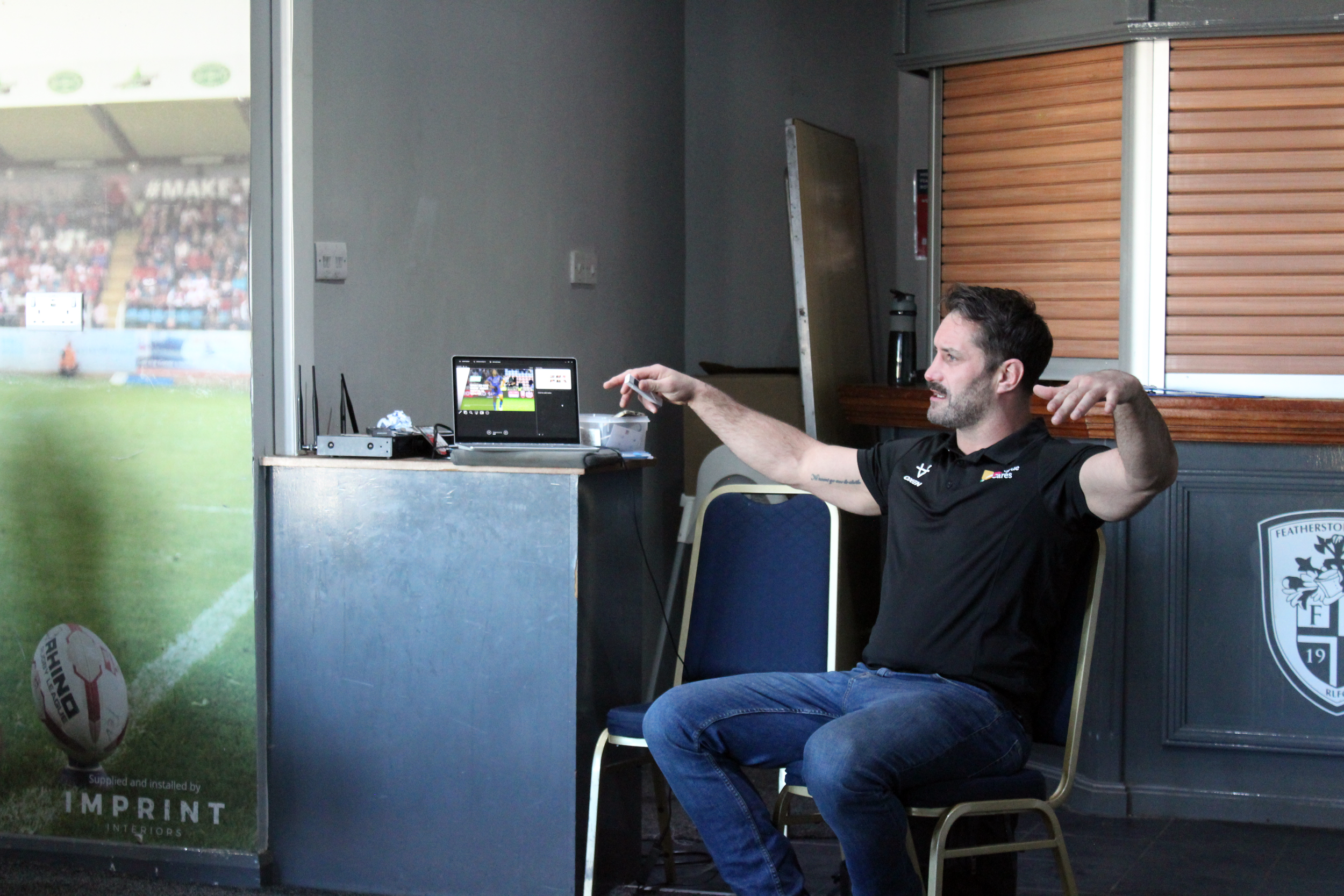 A man sitting on a chair inside a room, pointing with his right hand towards a laptop on a table. The laptop displays a cricket match. There is a rugby ball on the floor near the window, which shows a blurred view of a stadium filled with spectators.