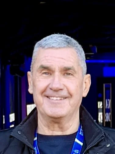 Smiling man with short gray hair, wearing a black jacket and a lanyard, standing indoors.