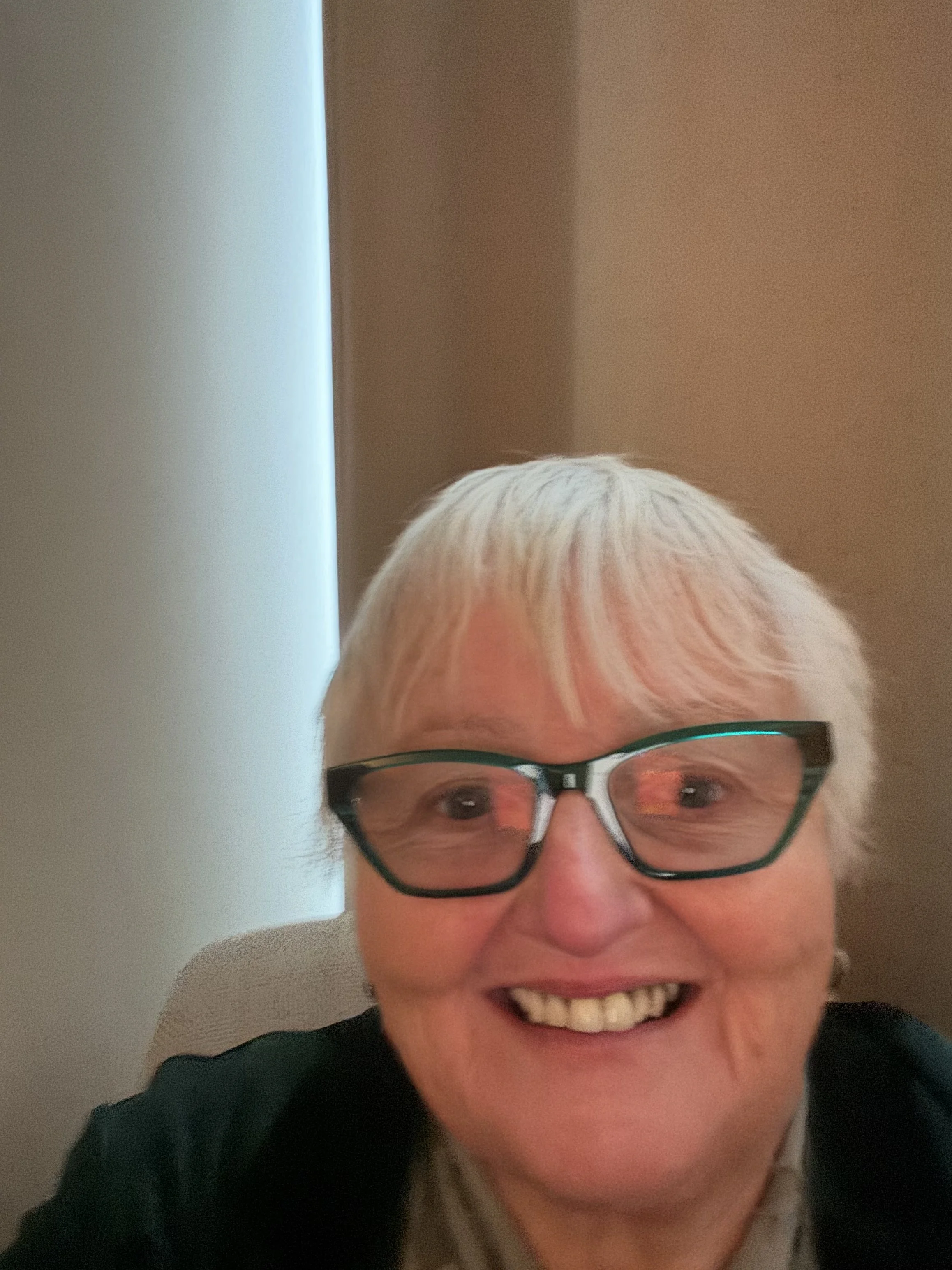 A smiling woman with short white hair, wearing glasses, is looking directly at the camera. The background shows a plain wall with a small section of a window or light source on the left.