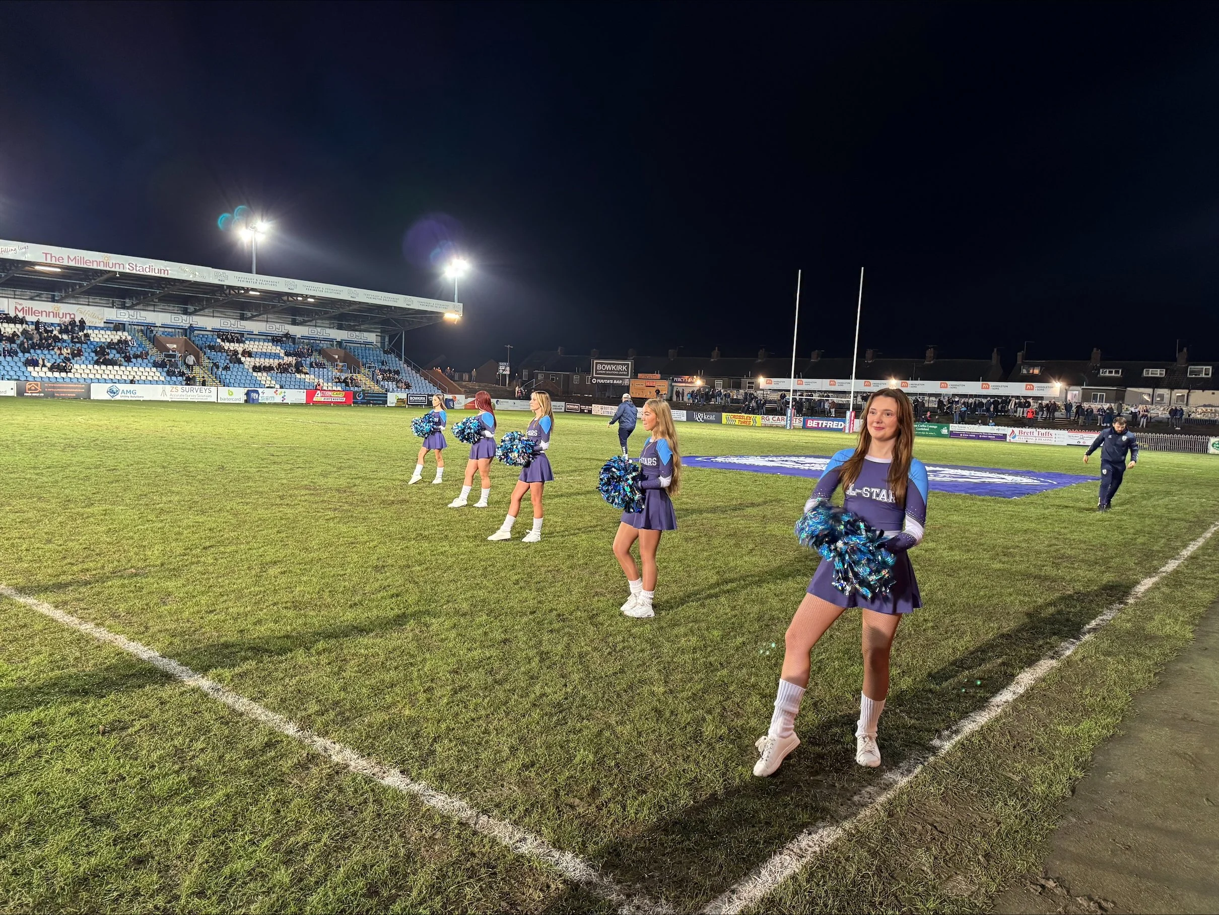 Cheerleaders standing on a football field at night, with a stadium crowd and empty goalposts in the background.