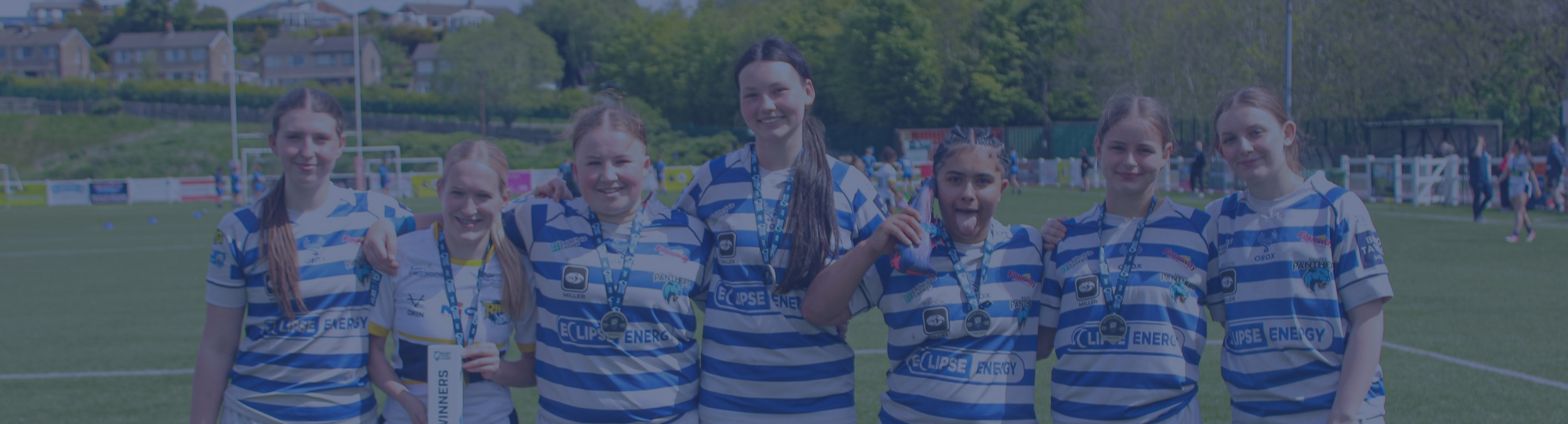 A group of seven young girls wearing blue and white striped sports jerseys, standing on a sports field after a match, smiling and showing medals.