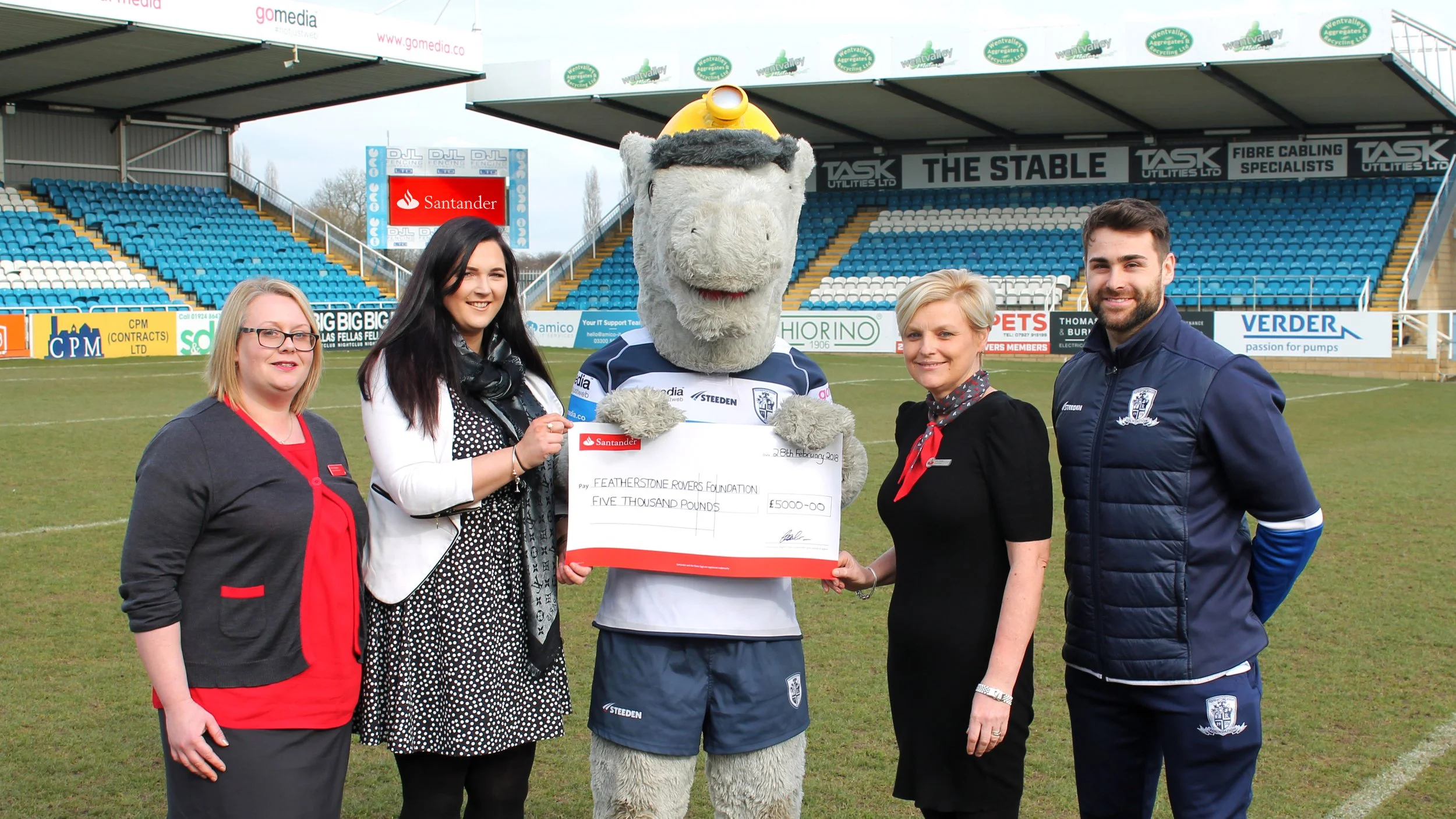 Five people and a large mascot holding a giant cheque at a sports stadium.