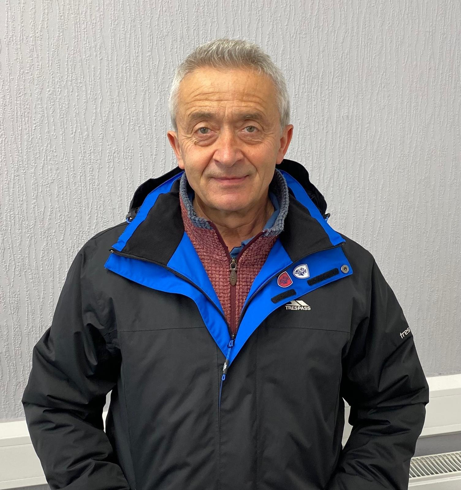 A smiling older man with gray hair, wearing a blue and black Trespass jacket over a red sweater, standing in front of a beige textured wall.