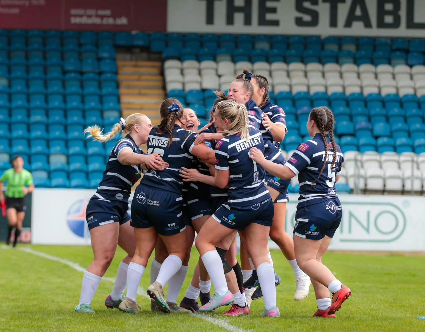 Women’s rugby team celebrating on the field, wearing navy blue jerseys and white socks, with some players hugging and smiling. The background shows stadium seats and a coach or referee in a green shirt.