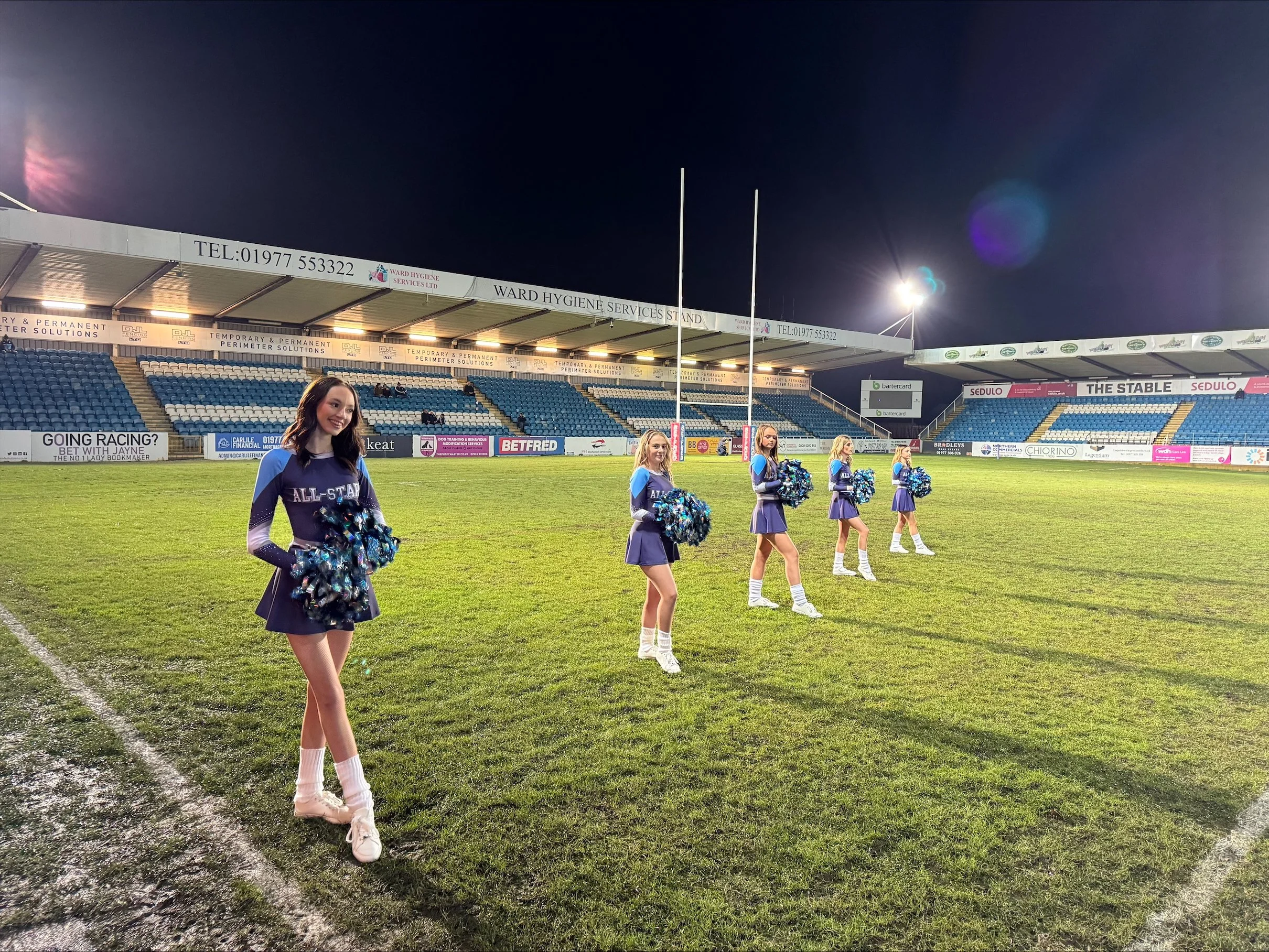 Cheerleaders standing on a sports field with floodlights, stadium seating, and advertising signs in the background during the night.