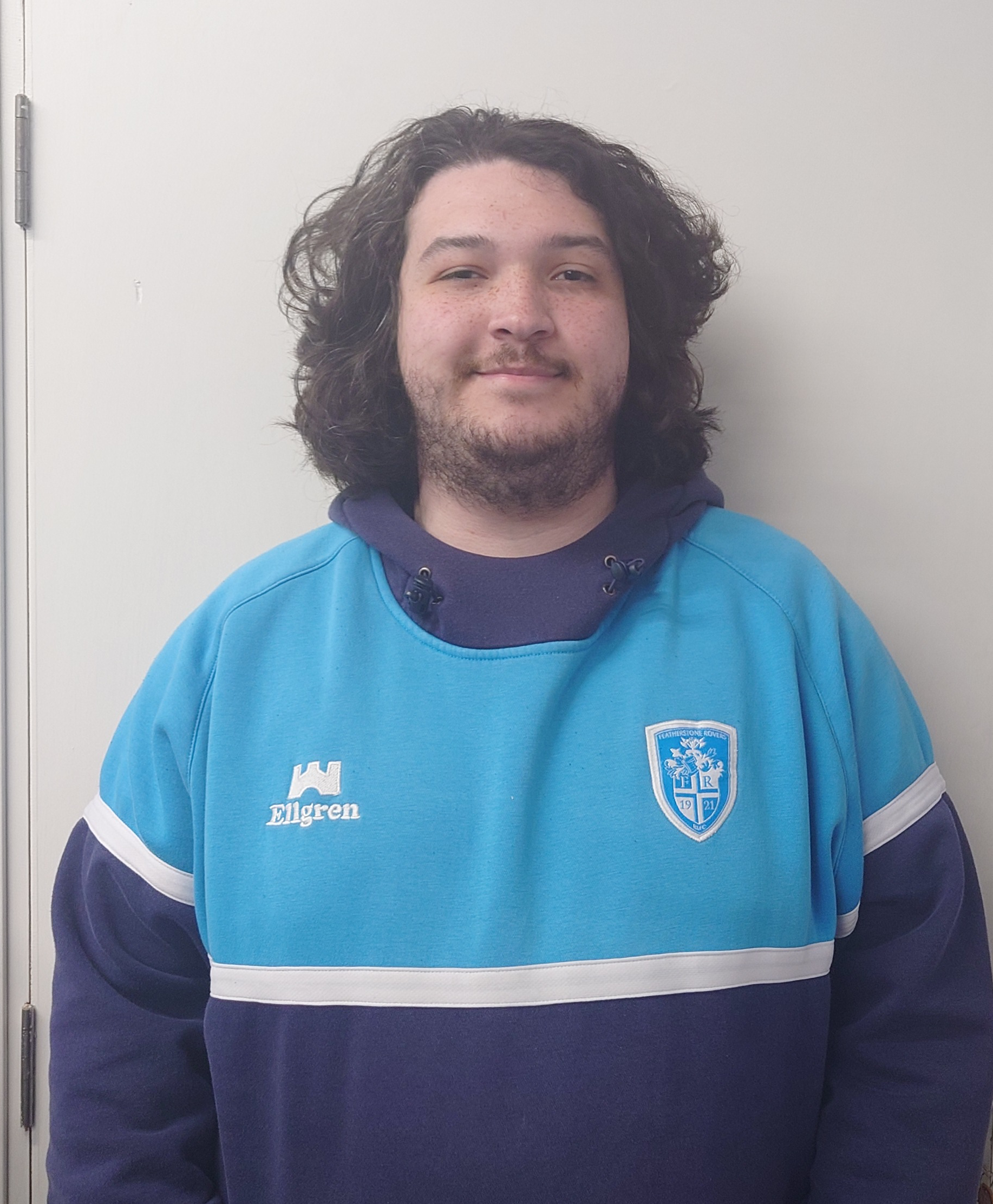 Close-up of a young man with long, curly dark hair, facial hair, and a slight smile, wearing a light blue sports jersey with a crest and the word 'Elgrens' on it, standing against a plain white wall.
