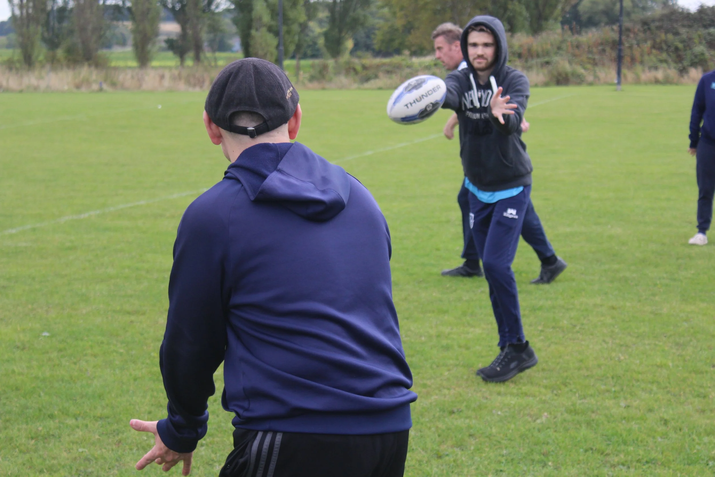A group of young men playing rugby outdoors on a grassy field, with trees in the background. One player is holding a rugby ball and appears to be passing or throwing it, while others are observing or preparing for the next move.