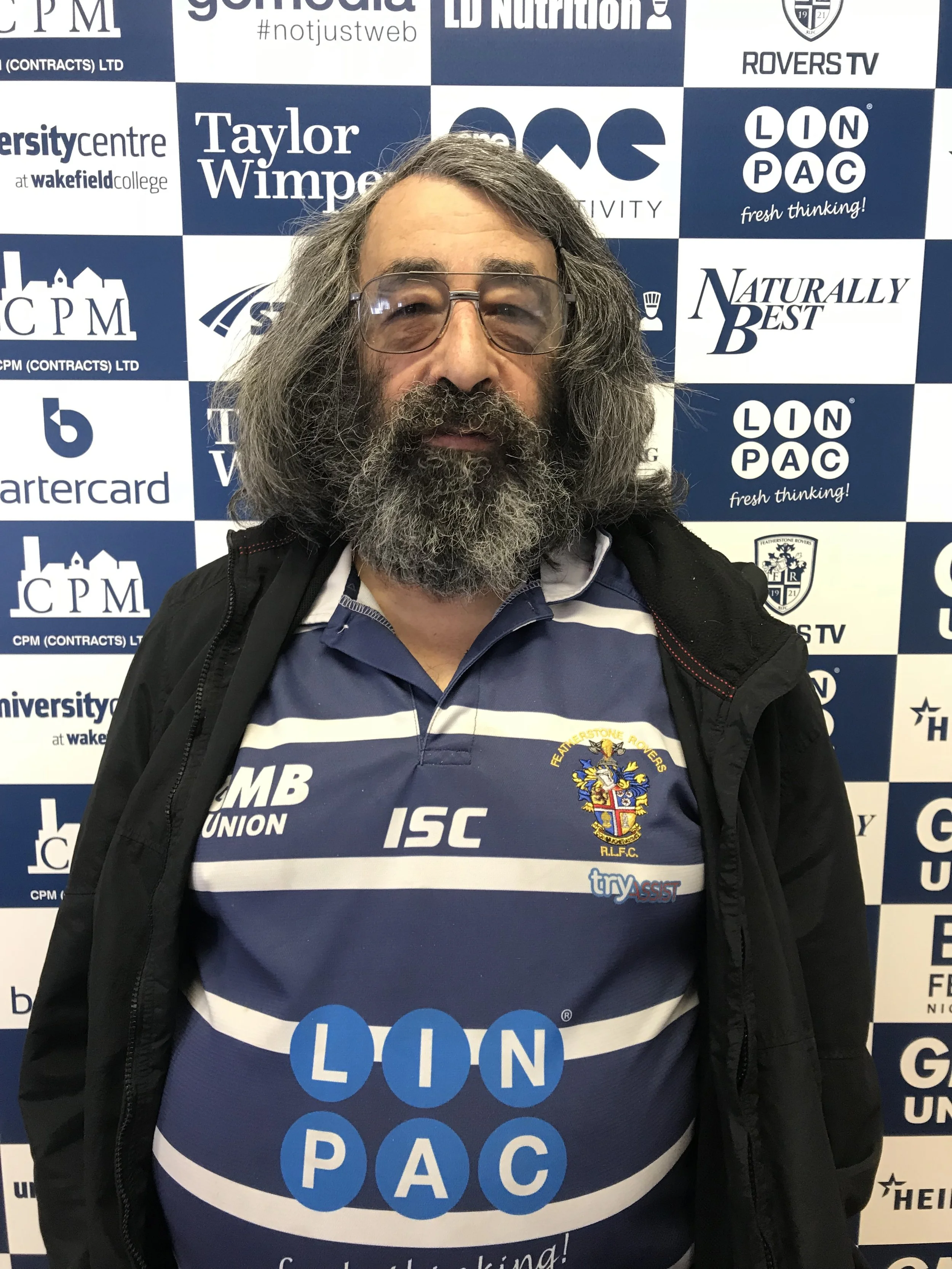 A man with gray, shoulder-length hair, a beard, and wearing glasses stands in front of a blue and white background covered with various sponsor logos, including 'LION PAC,' 'Taylor Wimpey,' 'Naturally Best,' and others. He is wearing a blue and white striped sports jersey with a crest and texting the word 'LION' across the front.