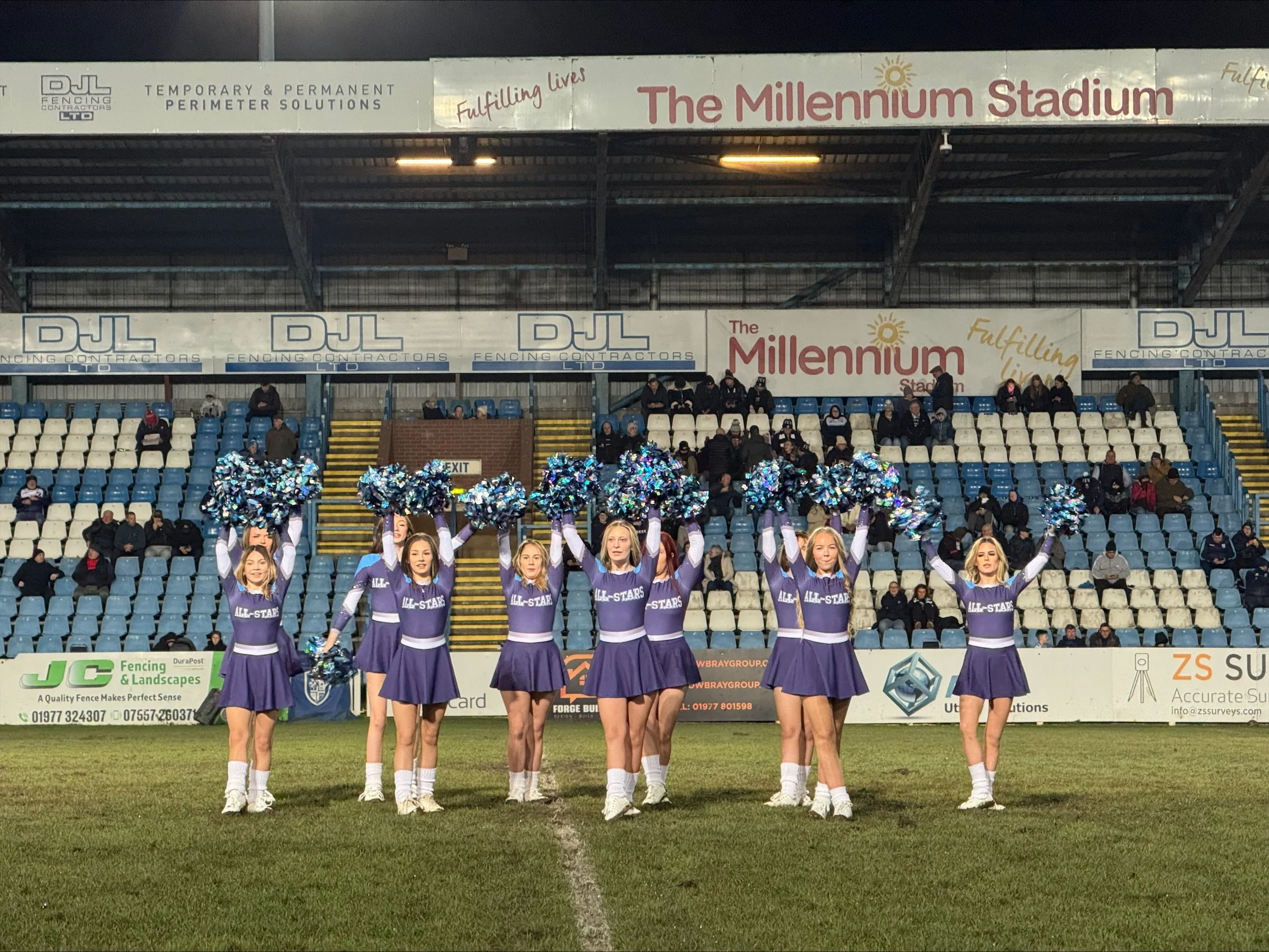 Cheerleaders in purple uniforms performing drills on a football field with spectators in the background and stadium signage.