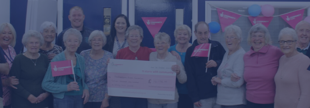 Group of smiling senior citizens holding a large check and pink flags at a community event.