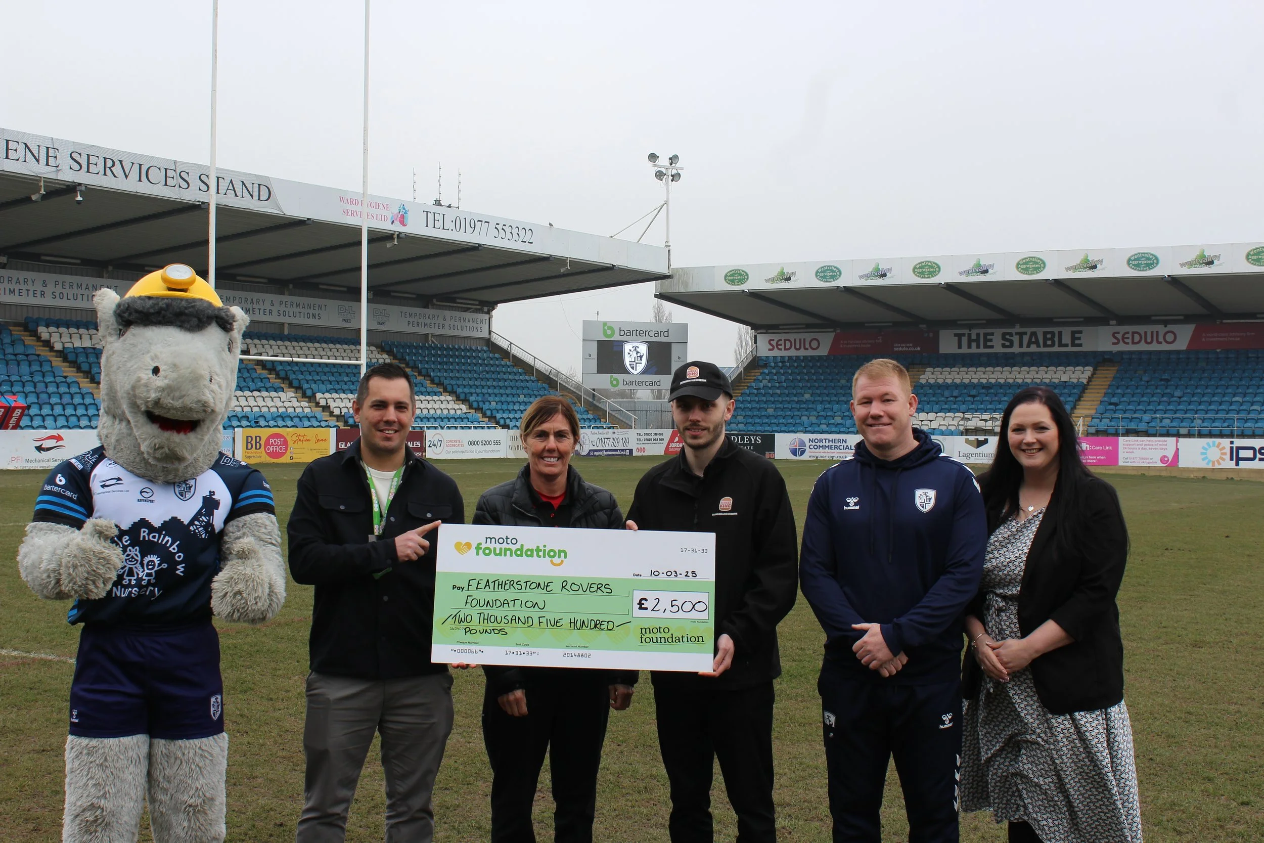 Group of five people and a mascot holding a large check on a sports field with stadium seats in the background.