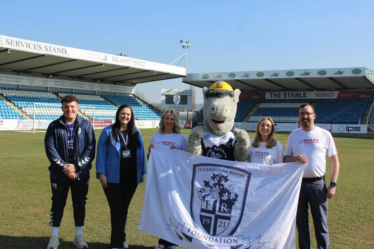 Group of five people and a mascot standing on a football field holding a Featherstone Rovers flag, with stadium seating in the background.
