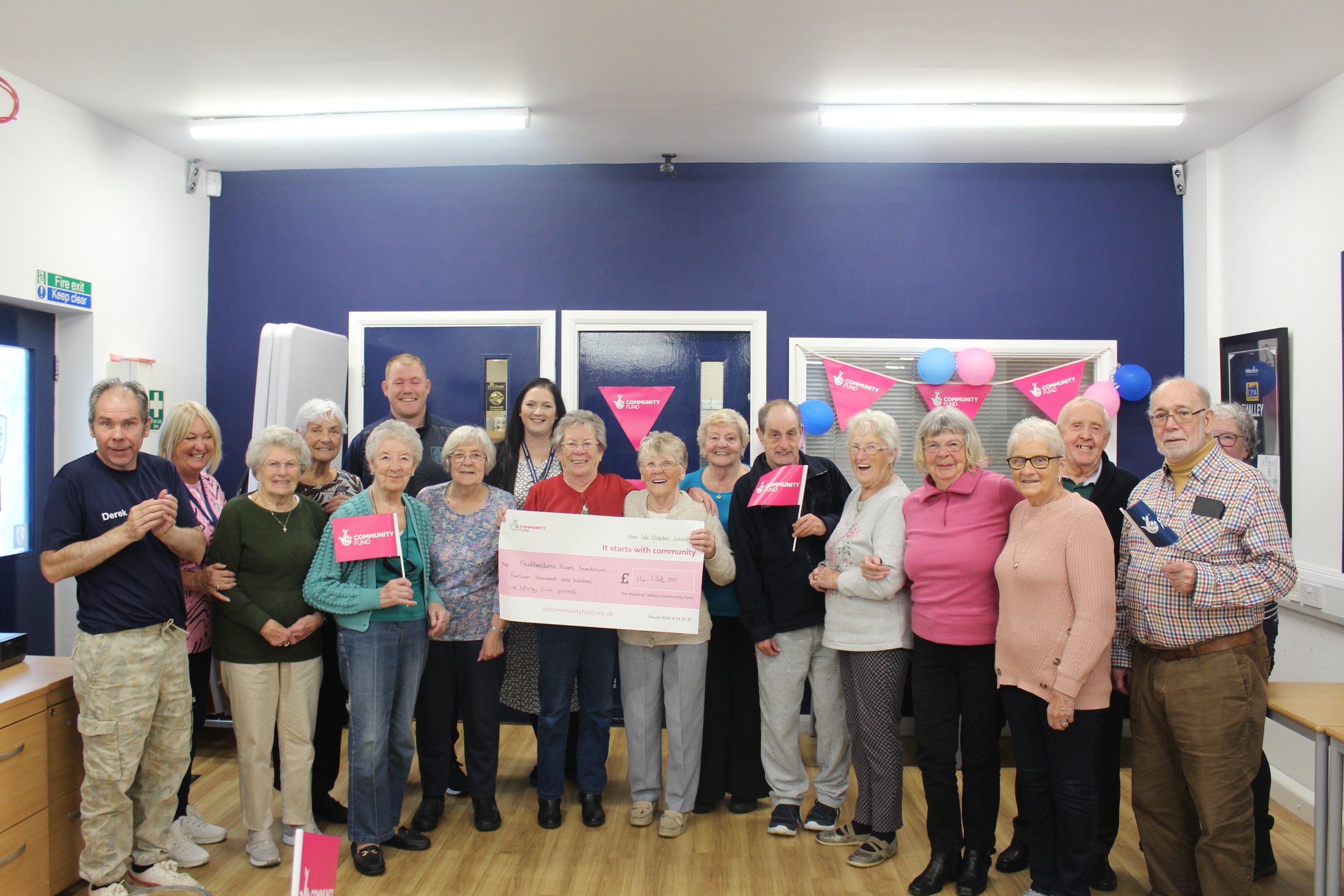 A group of elderly people and a few younger adults gathering indoors, holding pink flags with the words 'Community Fund' and a large check for over 14,000 pounds, celebrating a donation or grant. The background has blue and pink decorations, balloons, and signs indicating a charity event or community celebration.