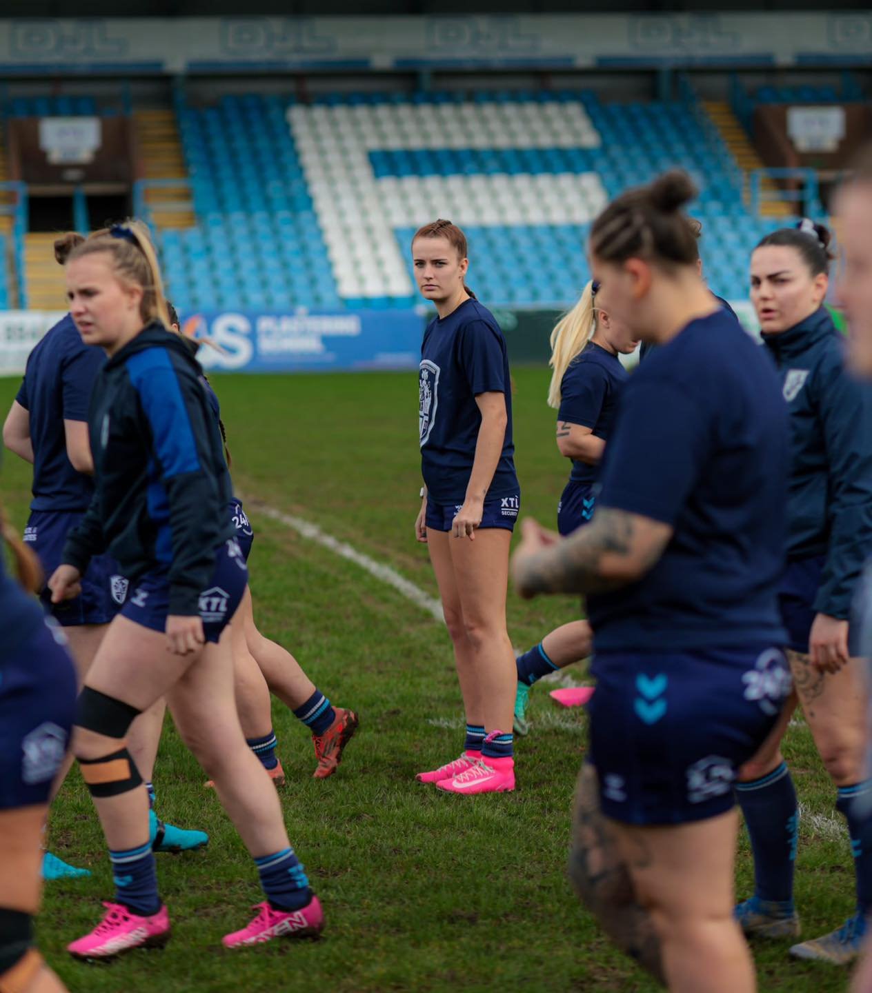 Australian female rugby players standing on the field during training or practice session, wearing blue team uniforms with some players with visible tattoos and others with ponytails, in a stadium with blue and white seating and playing field outside the stands.