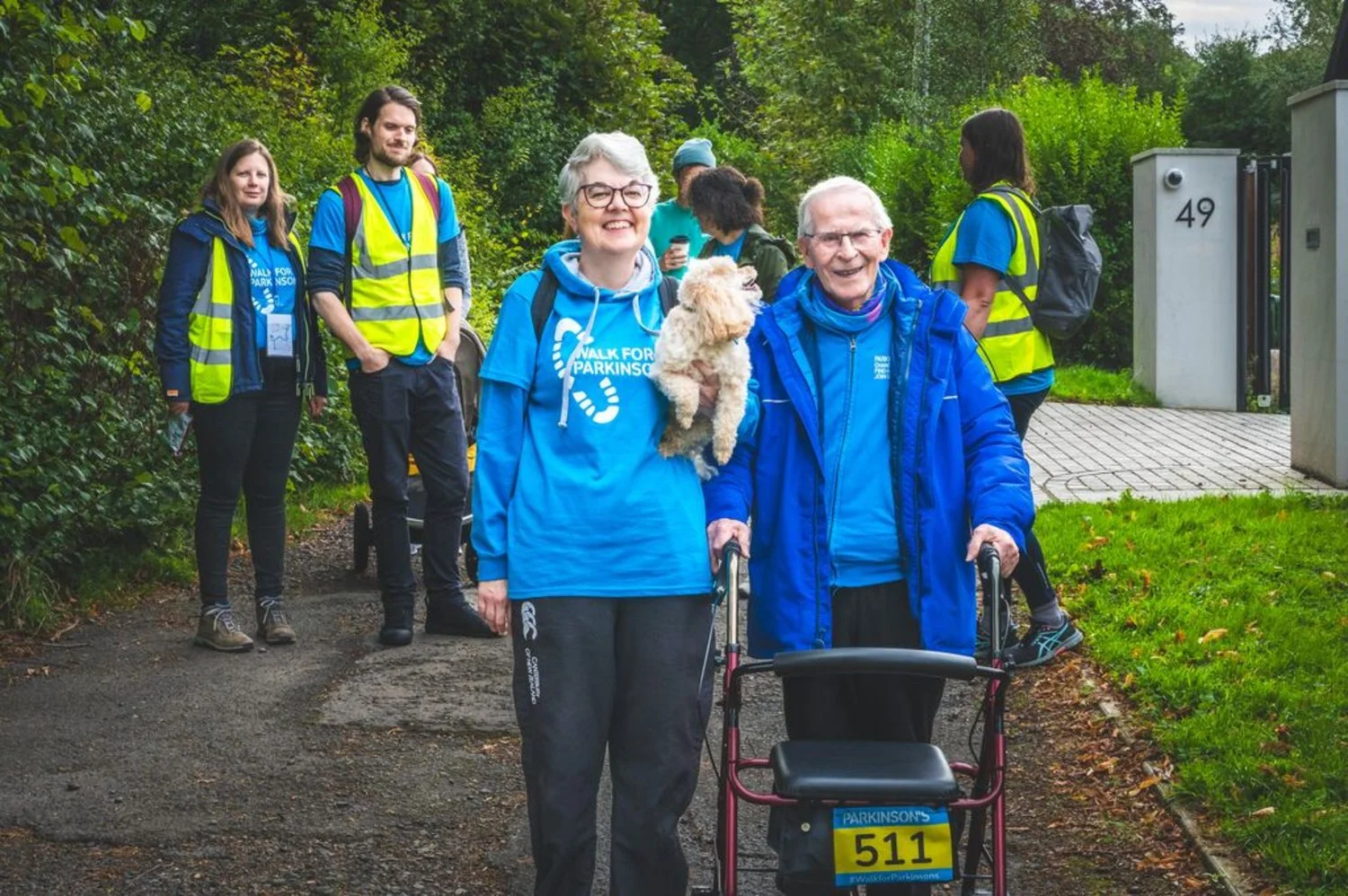 People participating in a walk for Parkinson's event outdoors, wearing blue shirts and yellow safety vests, with some using walkers and a dog present among the group.