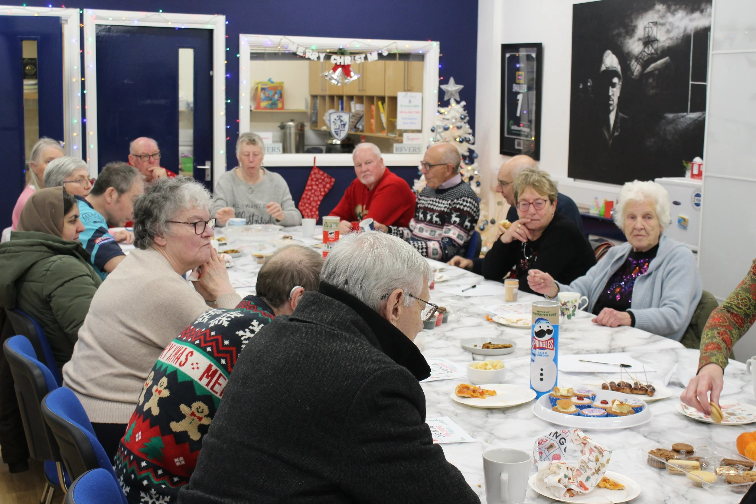 A group of elderly people gathered around a long party table decorated with Christmas decorations, eating and socializing in a room with holiday decorations including a small Christmas tree, lights, and festive banners.