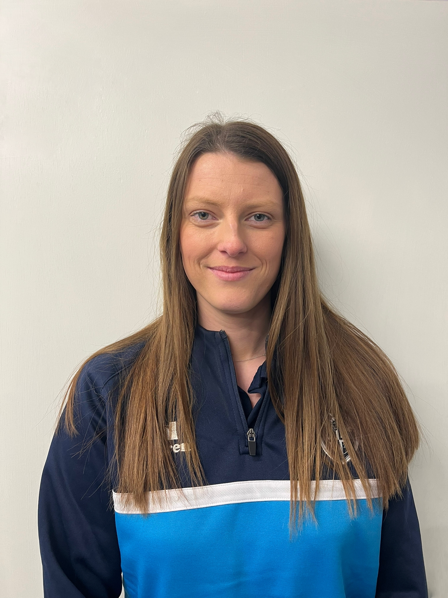 A woman with long brown hair and blue eyes wearing a dark sports jacket with a blue and white design, standing against a plain off-white wall.
