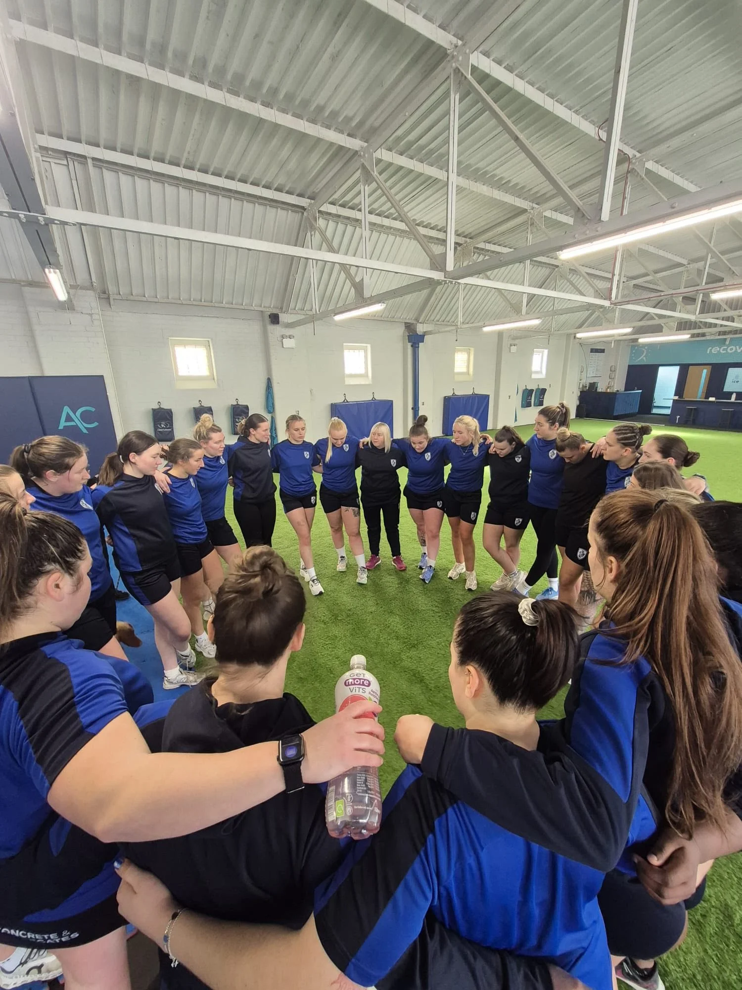A group of women in blue and black sportswear huddle together in a circle on an indoor sports field, likely for a team meeting or pep talk.