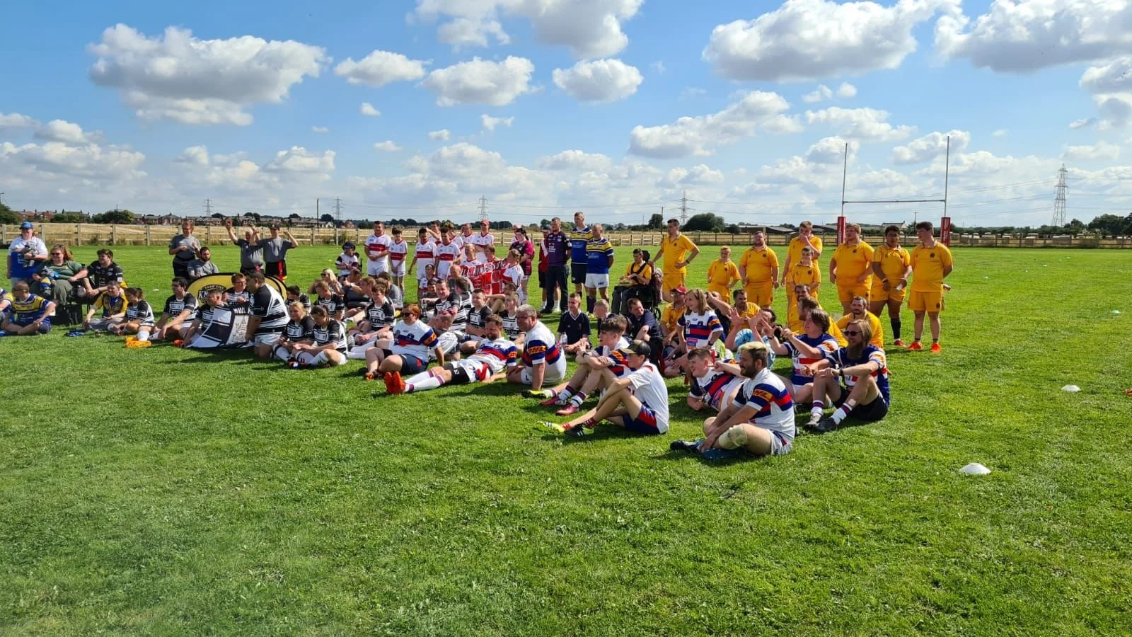 Group of rugby players and spectators on a grassy field under a blue sky with clouds, with players in yellow, purple, and striped uniforms.